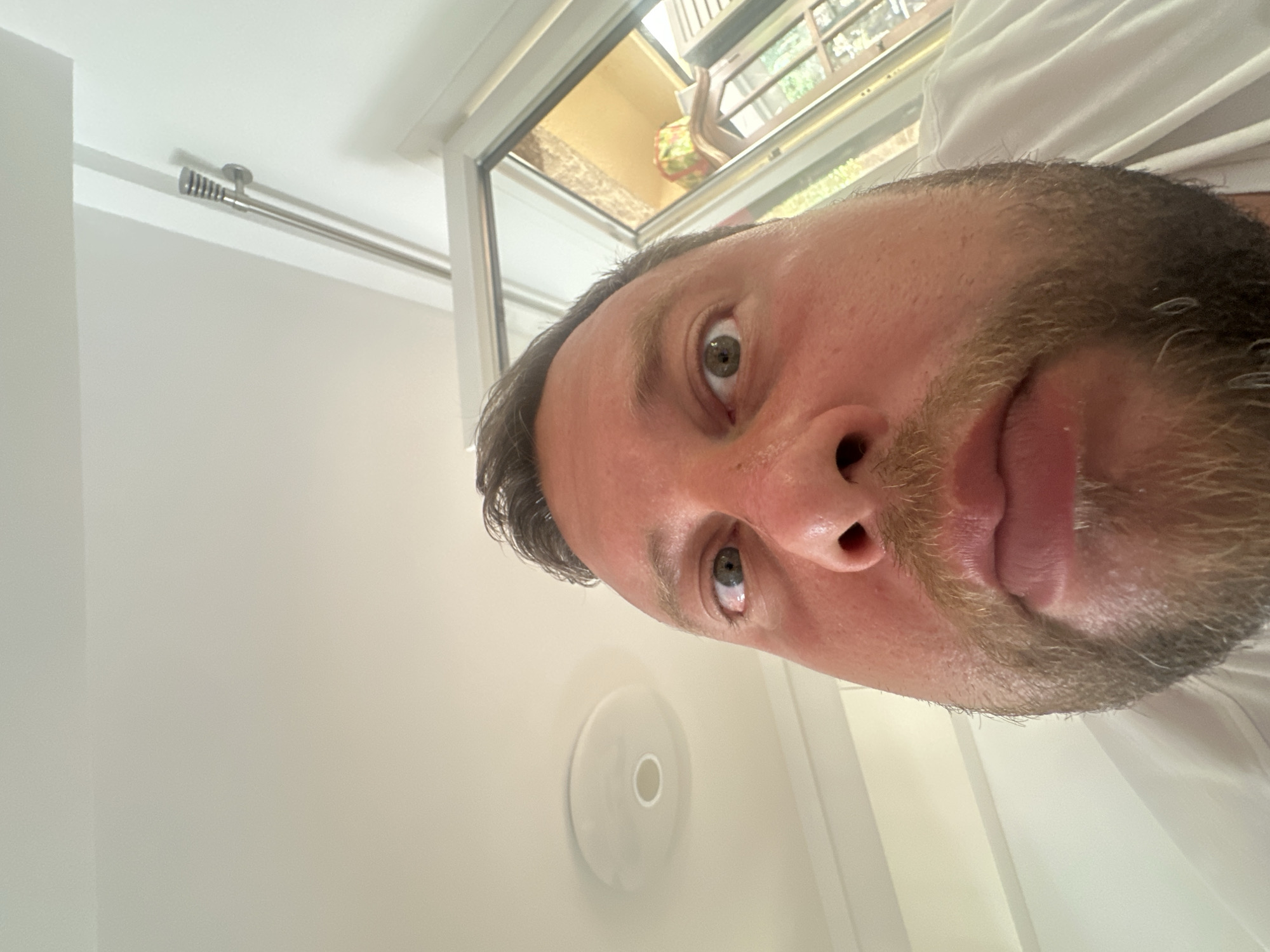 A low-angle, close-up selfie of a man's face fills the frame, shot from the chin upwards, with his eyes wide and looking directly at the camera. He has fair skin, reddish cheeks, brown hair, and a light brown beard. His mouth is slightly open, revealing his lips. Behind him, a white ceiling with a circular light fixture is visible. To the right, a window or glass door shows an outdoor scene with greenery and a yellow-toned interior. The lighting suggests it is daytime. The image was taken in the city of Sant Feliu de Guíxols, Spain, at a private residence or indoor space.