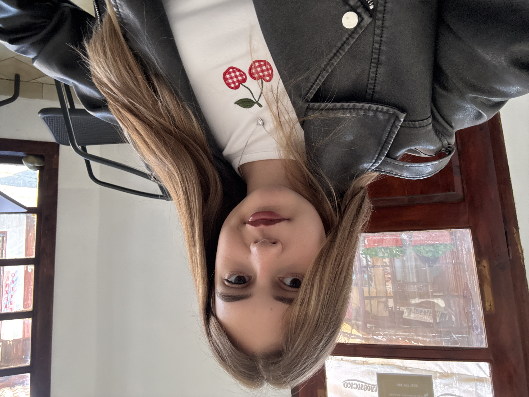 A portrait of a young woman, with light brown hair, wearing a white shirt with a cherry design and a black leather jacket. She is indoors, possibly in a cafe, near a window with a wooden frame. Reflections visible in the window suggest the location is in Panagyurishte, Bulgaria.