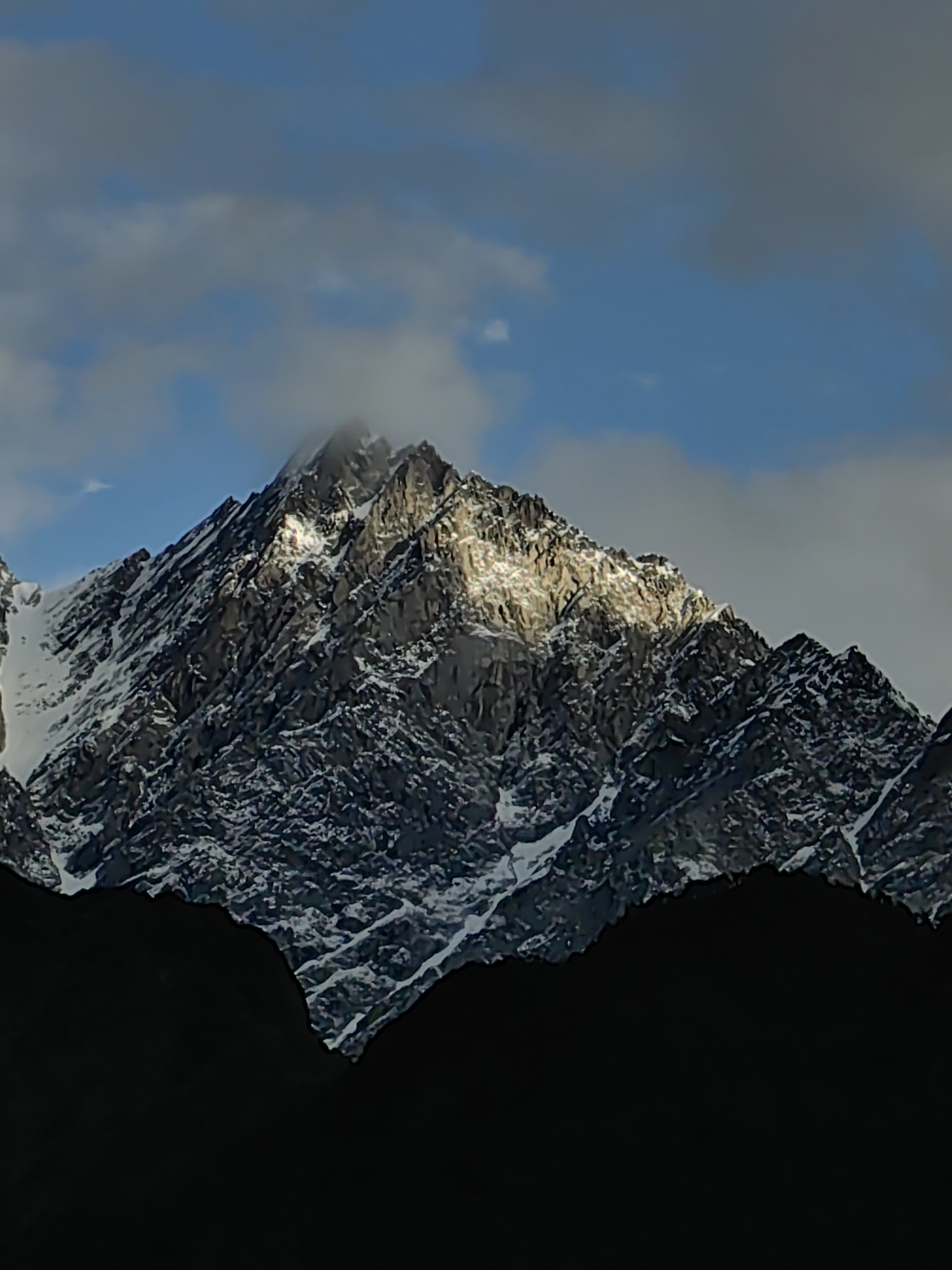 The image displays a majestic snow-capped mountain peak, partially illuminated by sunlight. The rugged terrain of the mountain is visible, with patches of snow contrasting against the dark rock. Clouds drift across a bright blue sky, with some areas appearing overcast. In the foreground, the silhouette of a darker mountain range or landmass creates a stark contrast against the illuminated peak. The overall scene is one of natural grandeur and the dramatic play of light and shadow on a mountainous landscape. The provided information indicates this scene is located in Vashisht, India.