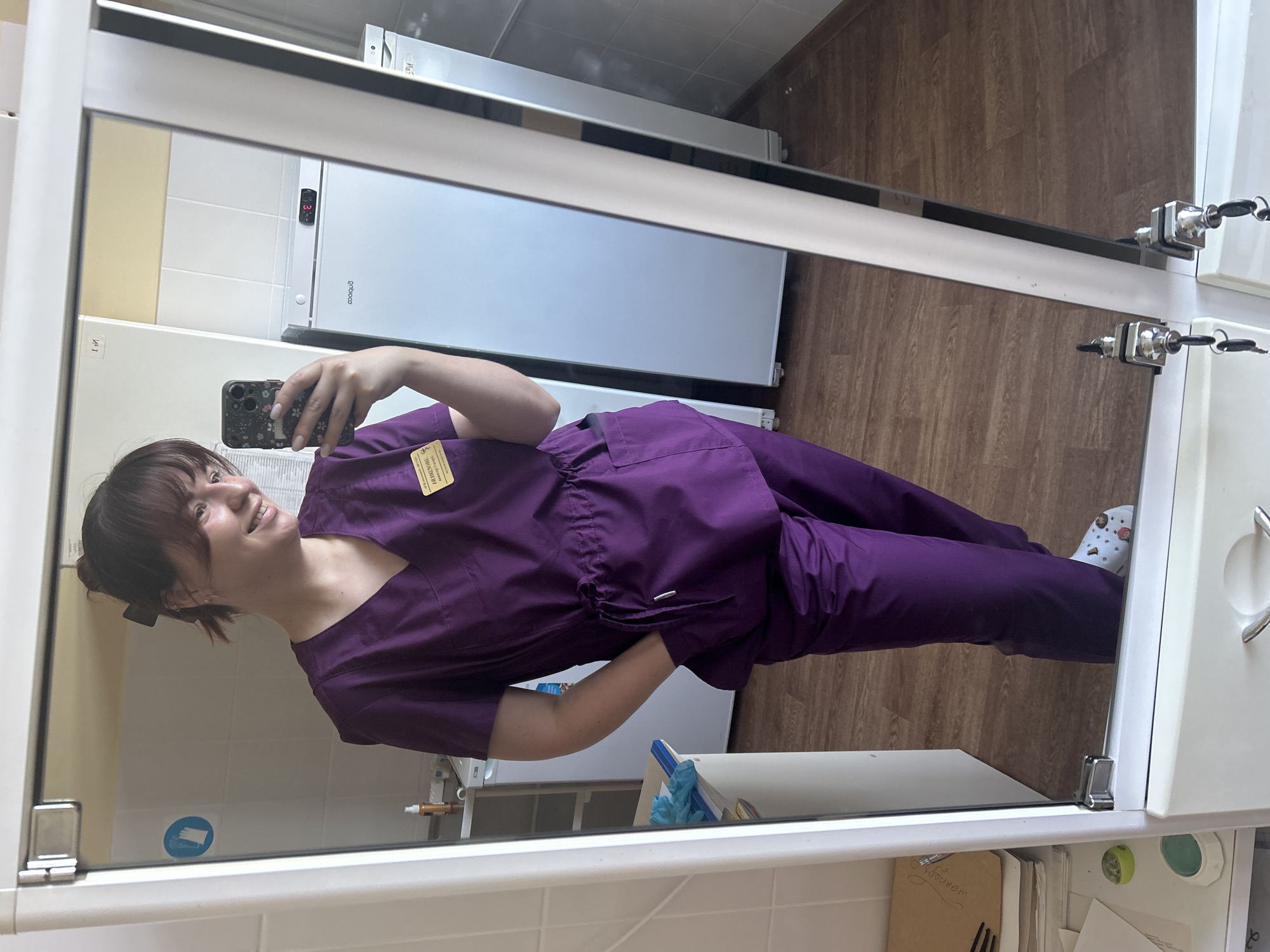 A young woman, appearing to be in her early twenties, is captured in a mirror selfie within what appears to be a medical or laboratory setting. She is wearing purple scrubs and has her hair pulled back in a bun with bangs. A yellow name tag is visible on her left chest. Her right hand is in her pocket, and she holds a smartphone in her left hand, using it to take the photo. The background reflected in the mirror shows tiled walls, two white refrigerators, and a wooden floor. On a shelf to the left, there are some medical supplies. There are no visible indications of the location being in the city of Фоки, Russia.