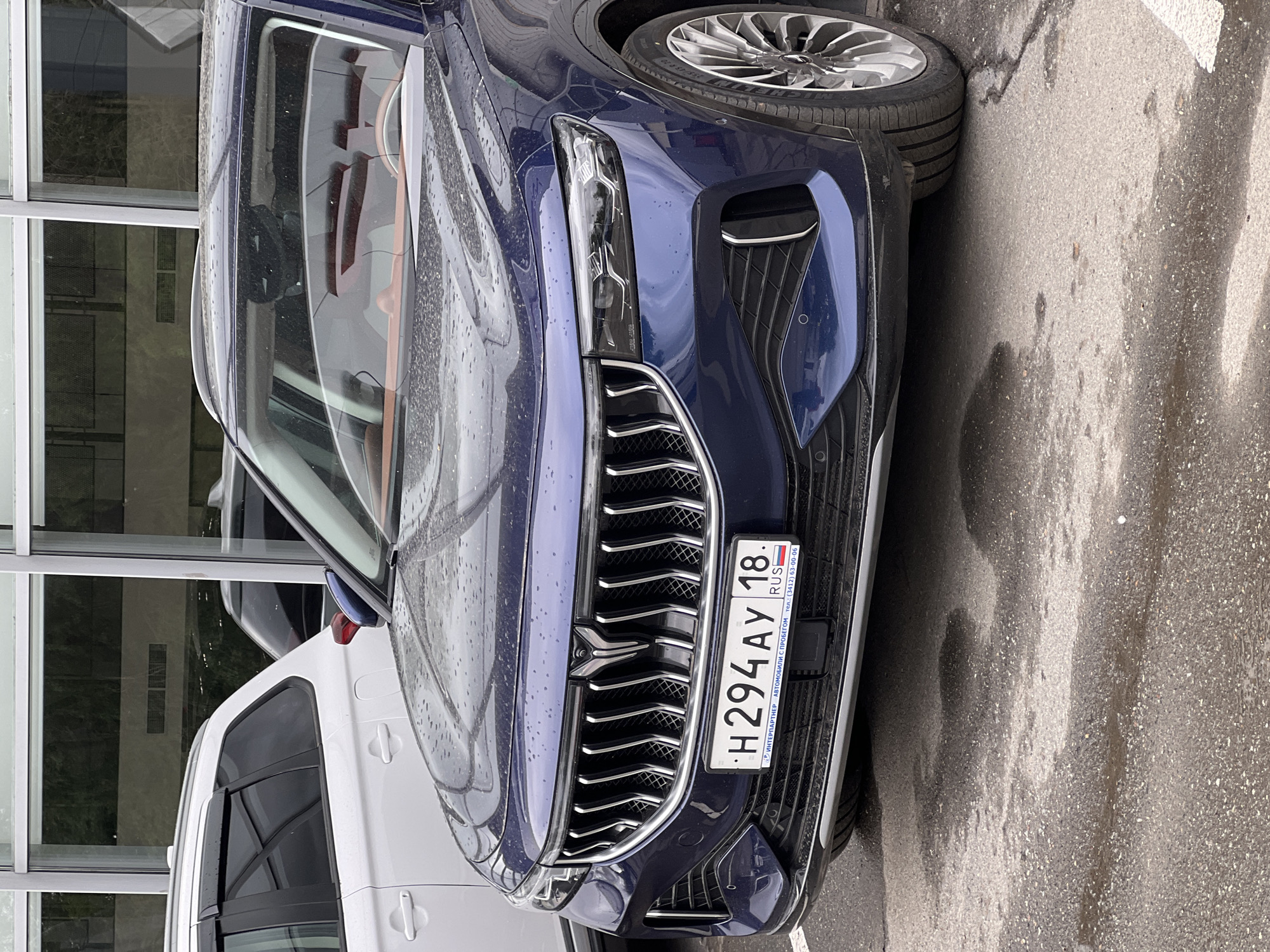 A dark blue Geely Monjaro (also known as Xingyue L) SUV is prominently parked on a wet asphalt surface, facing the viewer, in Izhevsk, Udmurt Republic, Russia. The vehicle features a large, vertically oriented chrome grille with a distinctive stylized "Y" emblem centered at the top. Flanking the grille are narrow, horizontally integrated LED headlights. Two circular parking sensors are visible on the lower front bumper on each side of the central intake. The Russian license plate, displaying `H 294 AY 18`, is mounted on a frame that reads "ИНТЕРПАРТНЕР АВТОМОБИЛИ С ПРОБЕГОМ тел.: (3412) 63-00-06", indicating the car is at an "Interpartner" dealership in Izhevsk (area code 3412, region 18 for Udmurt Republic). Through the windshield, brown leather interior elements, including the dashboard and seats, are discernible. The front right wheel has multi-spoke alloy rims, with the tire size 255/45 R20 101Y visible on the sidewall.
To the left and slightly behind the Geely Monjaro, a white third-generation Mitsubishi Outlander SUV is partially visible, showing its rear passenger side. This vehicle is equipped with roof rails and dark window deflector visors. The ground is a dark, wet asphalt parking lot, with numerous water droplets and small puddles reflecting the overcast sky, consistent with recent rainfall. In the background, a modern building with extensive glass windows and light-colored architectural frames is visible, suggesting a commercial or showroom environment. No people are present in the image.