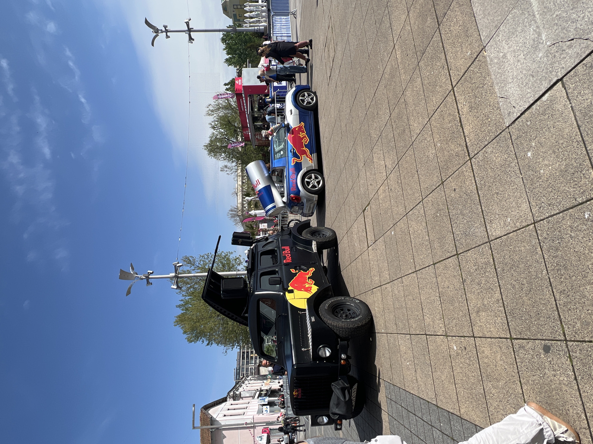 The image shows a city street scene in Burgas, Bulgaria, featuring two branded Red Bull vehicles parked on a paved surface.
In the foreground is a black, modified SUV-style vehicle with the Red Bull logo prominently displayed on its side. The vehicle has an open hood, revealing its engine components, and a tall, pole-like structure extending upwards from its roof, topped with a wind vane-like object.
Behind the black SUV is a smaller, blue and white car, also branded with the Red Bull logo. A large replica of a Red Bull can is mounted on the back of this car.
In the background, pedestrians can be seen walking along the street. There are buildings visible, including a light pink one. A lamppost and trees are also part of the background. A red banner with flags on the poles indicates the location is likely a public event or promotional area. The sky is blue with some clouds.