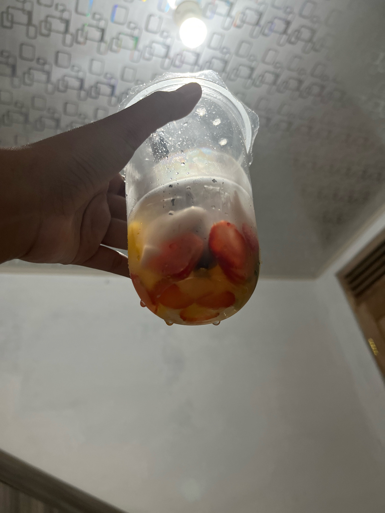 A hand holds a clear plastic cup filled with a beverage containing visible pieces of fruit, including strawberries and what appear to be peach slices. The cup is partially obscured by the hand, which is positioned in the lower left quadrant of the image. Water droplets are present on the exterior of the cup, suggesting it is cold. The background is out of focus but appears to be an indoor setting with a patterned ceiling and a light fixture. There are no visible identifying landmarks or clues that place this image specifically in Matesih, Indonesia.