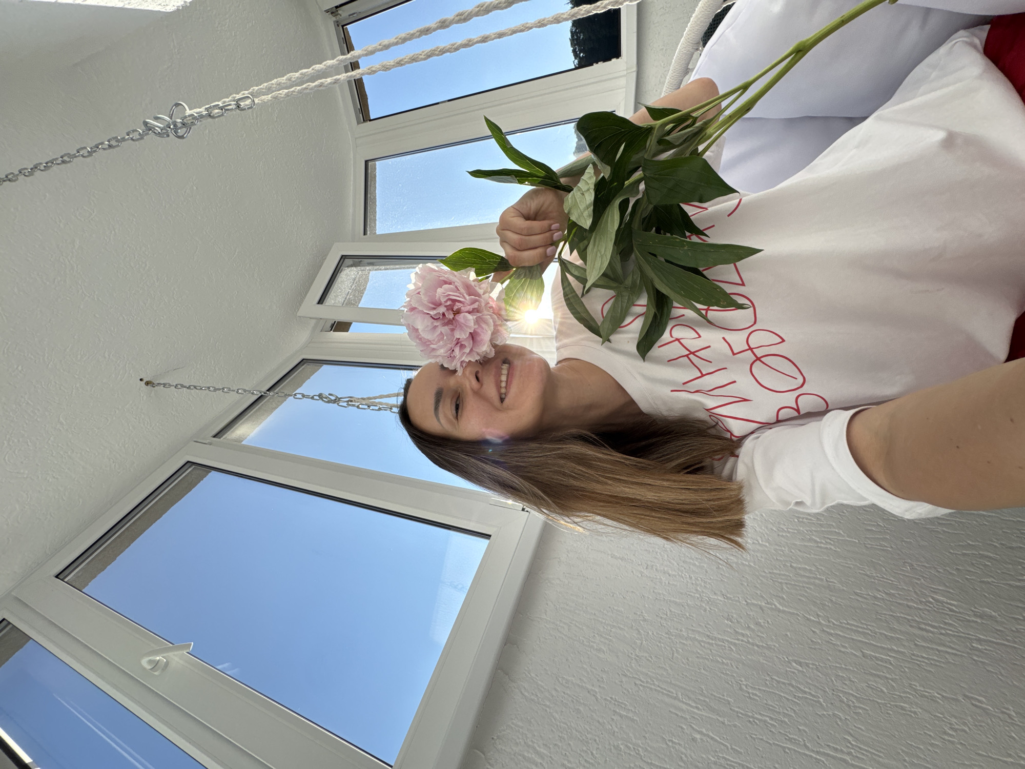A young woman with long brown hair is taking a selfie in a sunlit room with large windows. She is smiling and holding a pink peony flower up to her face, with its petals obscuring part of her face. She is wearing a white t-shirt with red lettering. The room appears to be a balcony or sunroom, with white walls and ceiling, and large windows offering a view of a clear blue sky. There are white chains hanging from the ceiling, suggesting a swing or hammock may be present. The bright sunlight creates lens flare and highlights the scene. The scene is likely within a residential building in Sochi, Russia, given the presence of a balcony and the sunny weather.