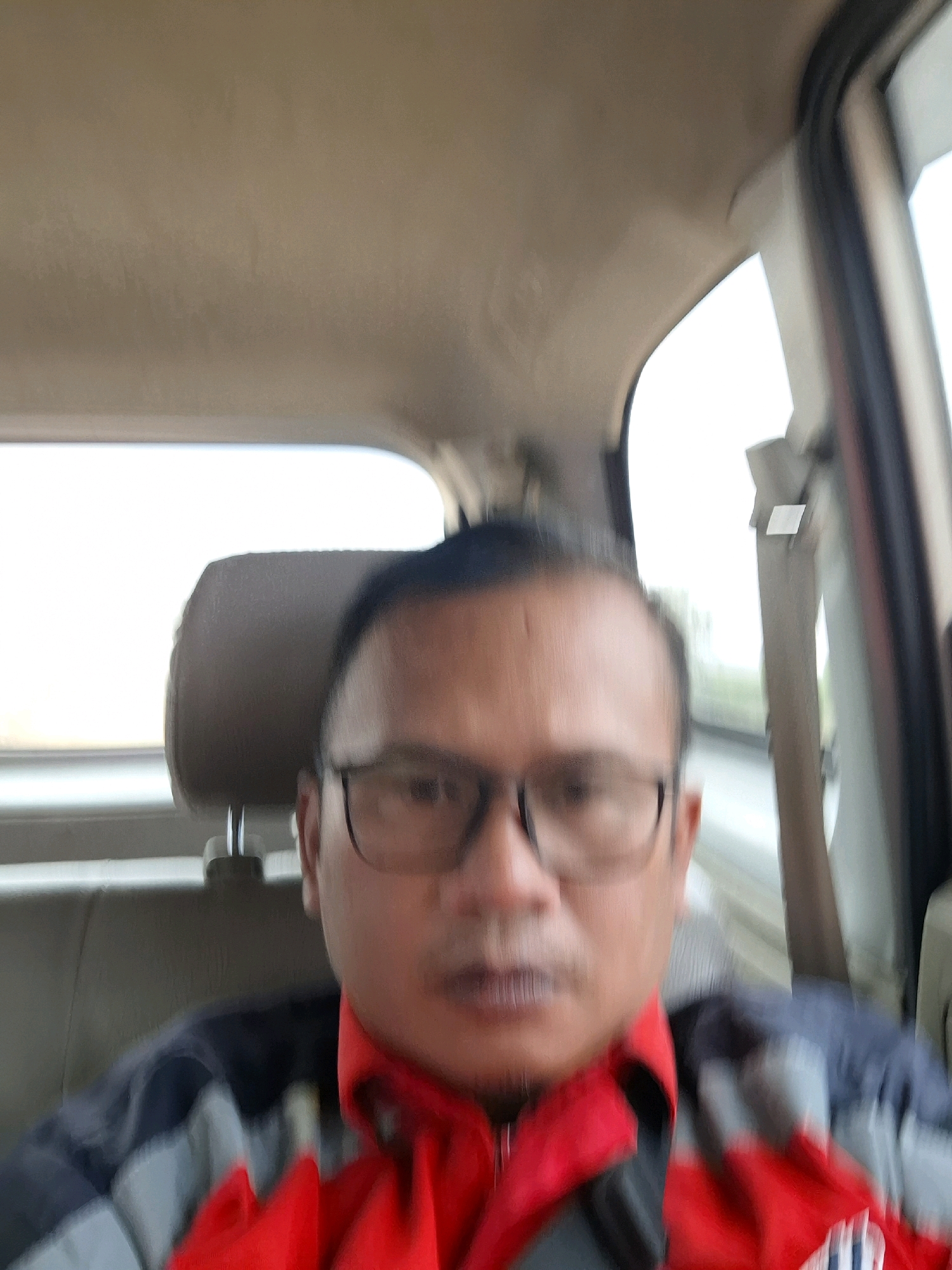 A man wearing glasses and a red collared shirt under a jacket is visible from the chest up, taking a selfie inside a vehicle. He is seated in the front passenger seat, with the headrest of the driver's seat behind him. The interior of the car, including the ceiling and the car door window frame, is visible. The man's facial features are slightly blurred. The background outside the car window is bright and appears to be a cloudy day with some greenery. The image was captured in Kutasirna, Indonesia.