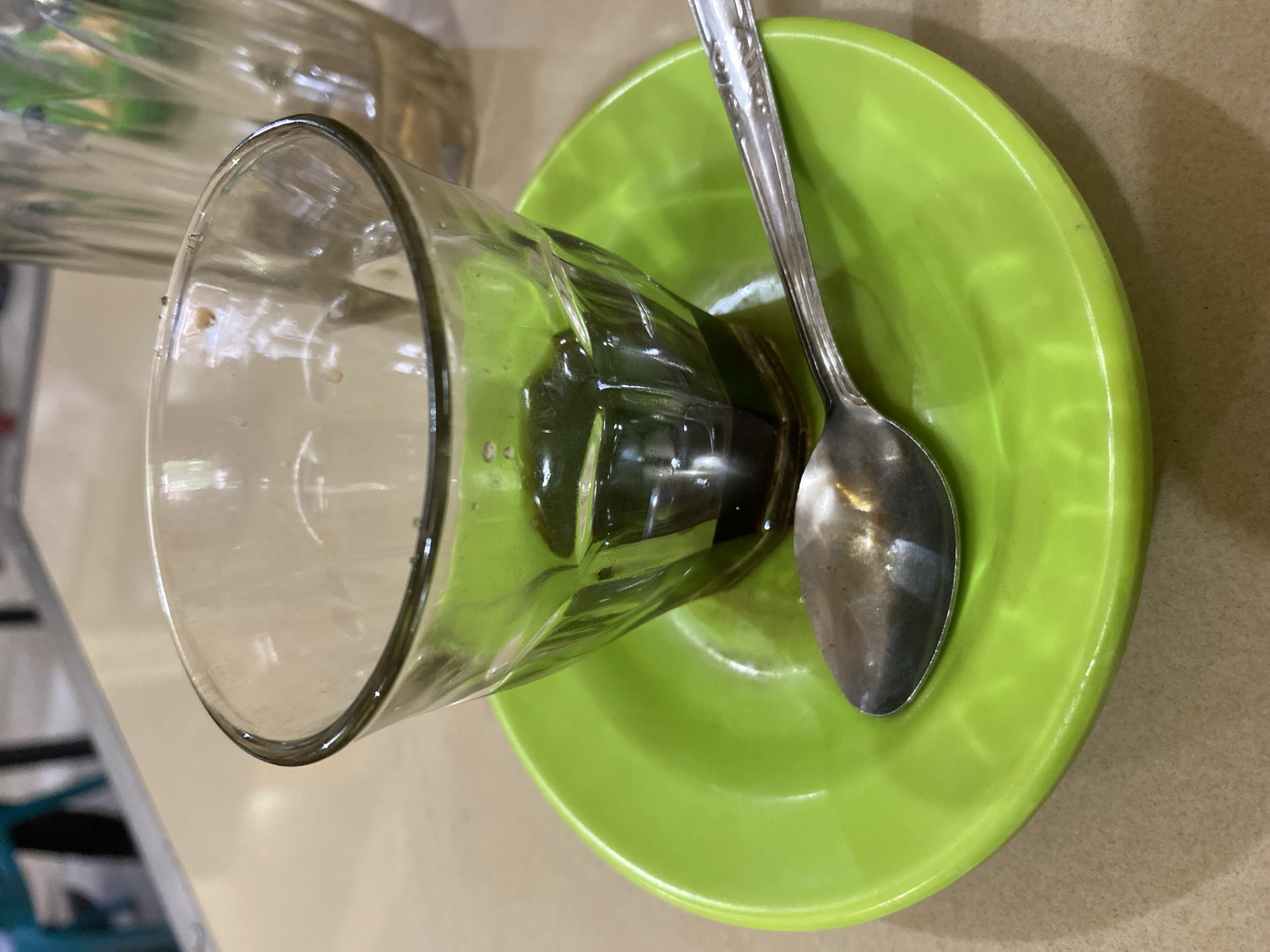 A close-up, eye-level shot captures a small, clear glass containing a dark liquid, likely coffee, resting on a bright green saucer. A metallic spoon, with an ornate design on its handle, lies on the saucer, positioned to the left of the glass. In the background, a partially visible glass pitcher and a blurred table surface are discernible, suggesting a casual dining or cafe setting. The scene is well-lit, emphasizing the textures and colors of the objects. This image was taken in Lhokseumawe, Indonesia, likely within a local establishment.