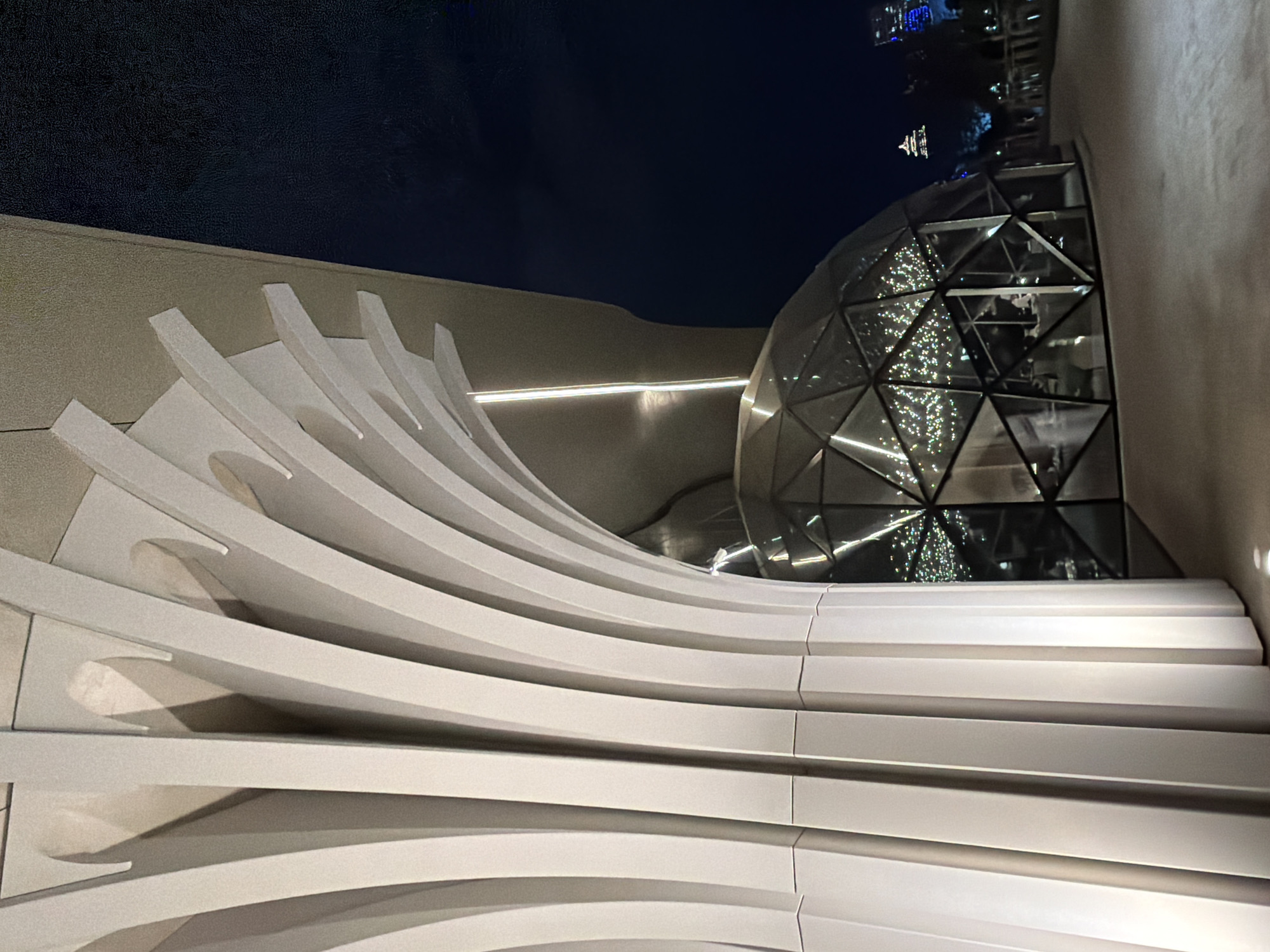The image depicts an outdoor scene at night. The primary subjects are the architectural elements of a modern building and the night sky. On the left, a series of white, curved fins create a dynamic, sculptural facade. These fins are evenly spaced and illuminated from within, casting shadows and highlighting their layered structure. To the right of the fins, a geodesic dome structure is visible. The dome is made of glass panels, reflecting the surrounding lights and the dark night sky. Inside the dome, string lights, likely Christmas lights, create a festive glow, illuminating the interior with scattered, bright points of light. In the distance, beyond the dome, faint lights of a city skyline are visible against the dark sky. There are no people or animals clearly visible in the image. The overall mood is sophisticated and modern, with a hint of festive cheer due to the lights within the dome. The time of day is clearly night, and the weather appears clear, allowing for a view of the distant city lights. The location is Astana, Kazakhstan, as indicated by the context provided. No text is visible in the image.