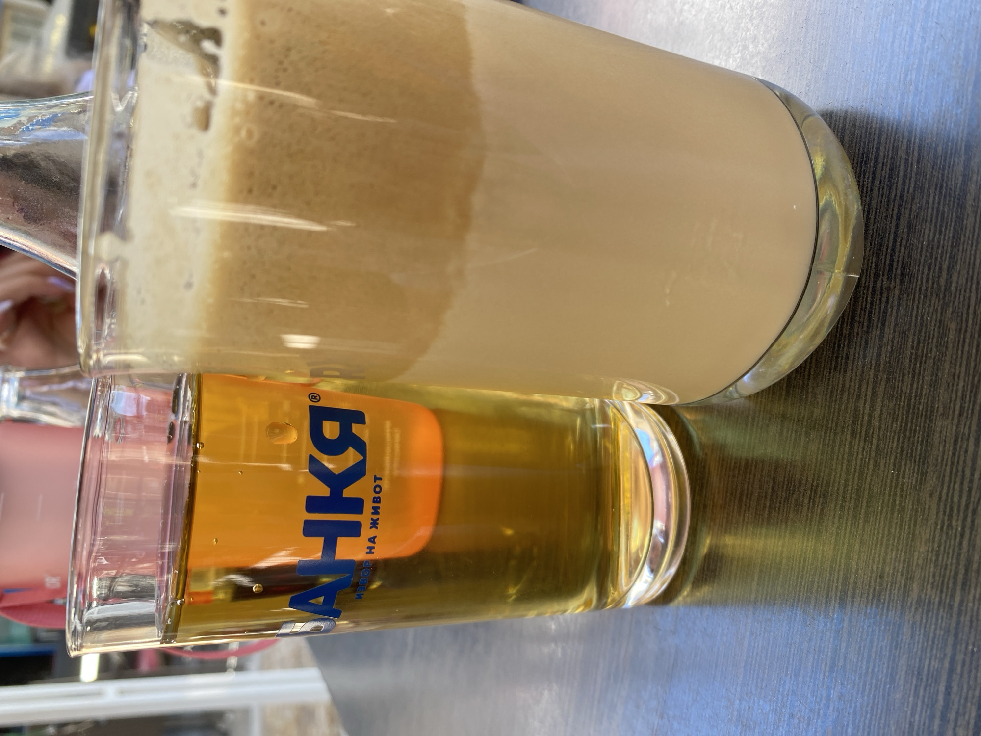 The image depicts two tall glasses on a dark wooden table, likely outdoors given the lighting and reflections. The glass on the left contains a golden-amber liquid, possibly beer or juice, with a small orange object visible inside. The glass on the right holds a layered beverage, with a lighter, frothy layer on top and a cream-colored liquid below. A pink backpack is visible in the blurred background, along with part of a white bottle. The text "БАНКЯ" and "ИЗВОР НА ЖИВОТ" is visible on the left glass. This appears to be a scene from Smolyan, Bulgaria, where Bankya mineral water is sourced.