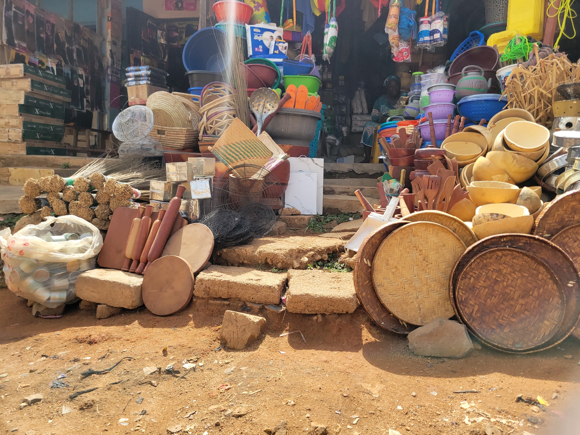 The image depicts an outdoor market scene in Jos, Nigeria. A variety of goods are displayed for sale, including woven baskets, wooden bowls and utensils, plastic basins, brooms, metal fans, and rolling pins. A woman sits in the background, likely tending the stall. The goods are arranged on and around concrete steps and the surrounding dirt ground. The area appears to be a bustling commercial zone.