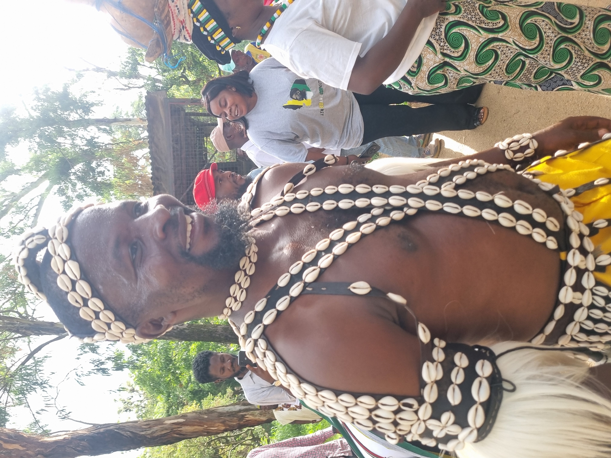 In Jos, Nigeria, a man dressed in traditional attire, adorned with cowrie shells around his head, neck, and arms, smiles at the camera. He wears a black leather harness embellished with numerous cowrie shells. A white, furry material is visible on his left arm. Behind him, a group of people are gathered. A woman in a grey t-shirt with a map graphic and text, and another woman in a white t-shirt with a beaded necklace, are in the foreground to the right. Other individuals are visible in the background, some wearing hats. The setting appears to be outdoors with trees and foliage. The ground is a light brown, dusty surface. Sunlight streams from the upper right of the frame, creating lens flare.