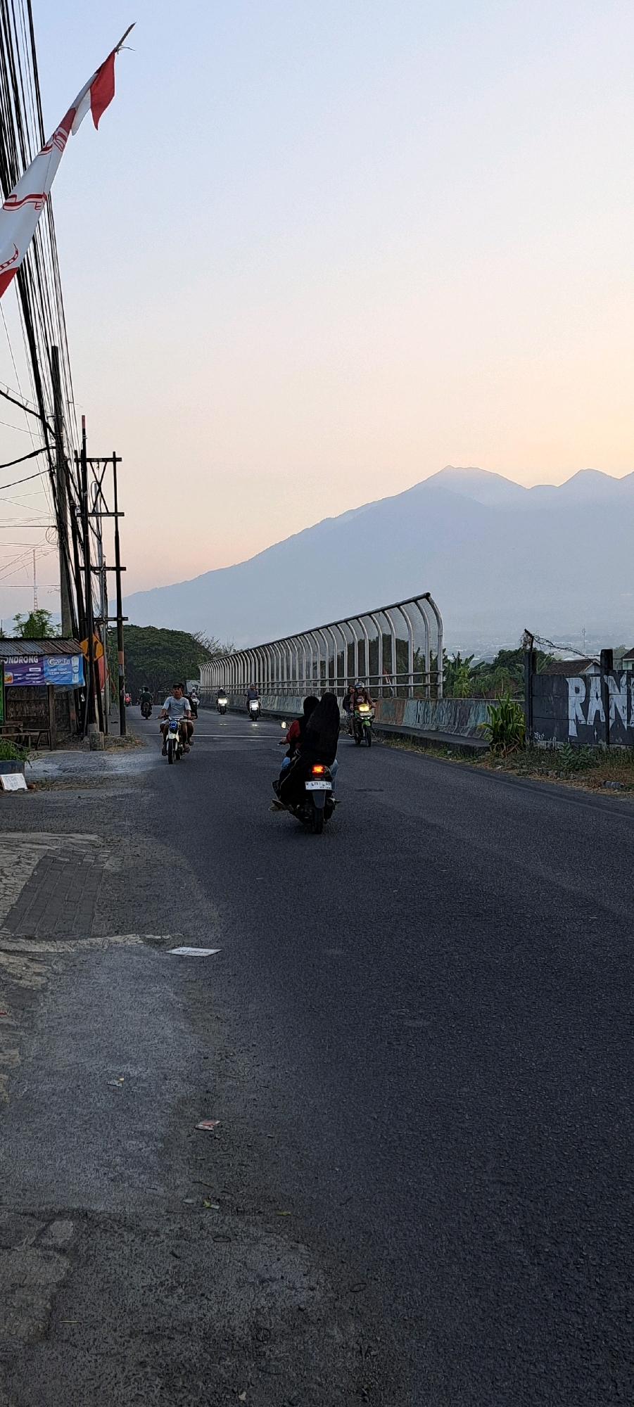 This image depicts a street scene in Randupitu, Indonesia, characterized by a road with several motorcycles in motion. In the foreground, a black motorcycle with two occupants, one wearing a headscarf, travels away from the camera. To the left, a motorcyclist wearing a light-colored shirt is seen approaching. Further up the road, other motorcycles are visible, indicating traffic. The road itself is asphalt and appears to be slightly wet in places.
On the left side of the road, there are utility poles with numerous wires and a small building with signage, one of which appears to be for "PONDRONG." A red and white flag, possibly the Indonesian flag, is fluttering on a pole.
The road is bordered on the right by a white metal barrier with a fence-like structure, suggesting it may be an overpass or elevated road. Beyond this barrier, vegetation and some buildings are visible. In the background, a large, hazy mountain range dominates the horizon under a pale sky with hints of sunset colors. The overall impression is a view of a developing area with infrastructure and natural landscape elements.