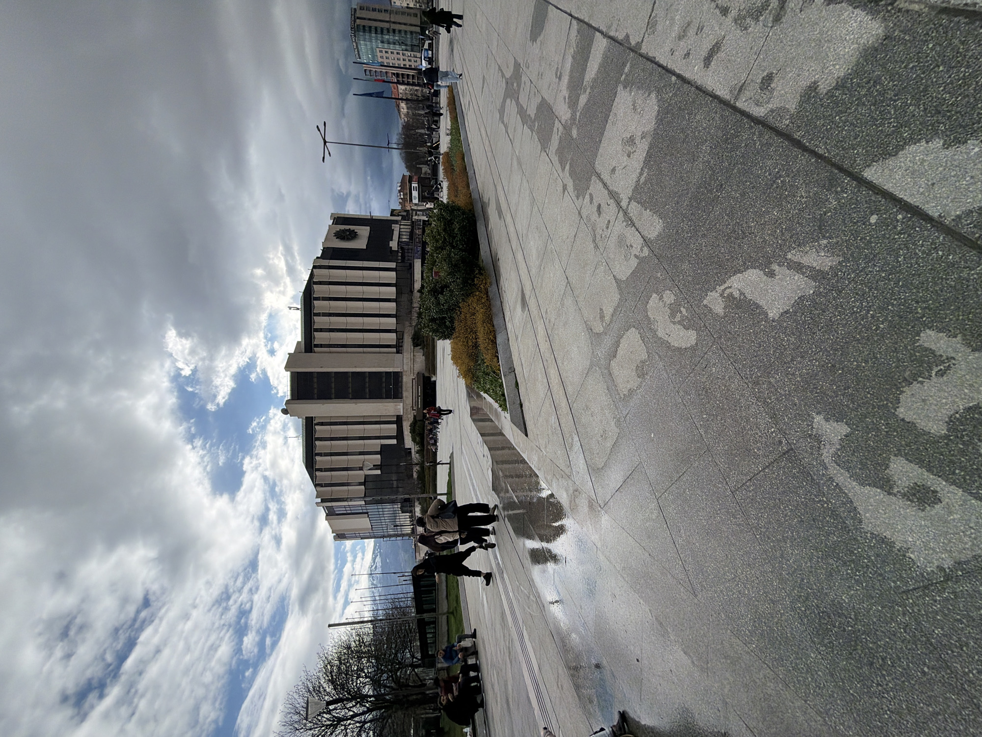 The image depicts a street scene in Sofia, Bulgaria, with the National Palace of Culture (NDK) prominently visible. The foreground shows a paved walkway with scattered puddles reflecting the sky. A group of people is walking along the walkway. In the background, other buildings and flagpoles are visible. The sky is cloudy with patches of blue.
