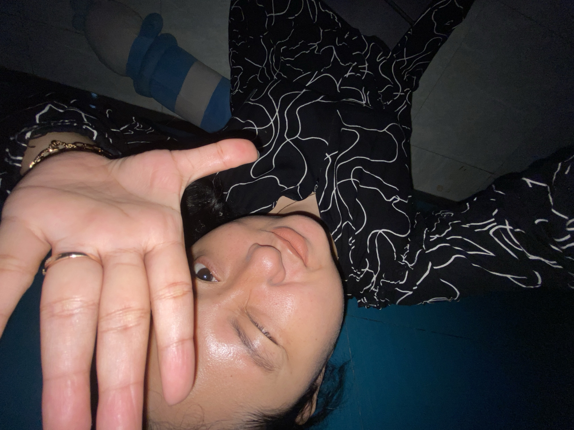 The image shows a person lying down on a blue surface, possibly a bed or floor, with a black and white patterned garment. The person is holding their hand up towards the camera, obscuring part of their face. A ring is visible on one of the fingers, and a bracelet adorns the wrist. In the background, a blue and white striped object, possibly a pillow or stuffed toy, is visible. The lighting suggests it might be evening or indoors. The overall mood is casual and relaxed. There is no visible text in the image.