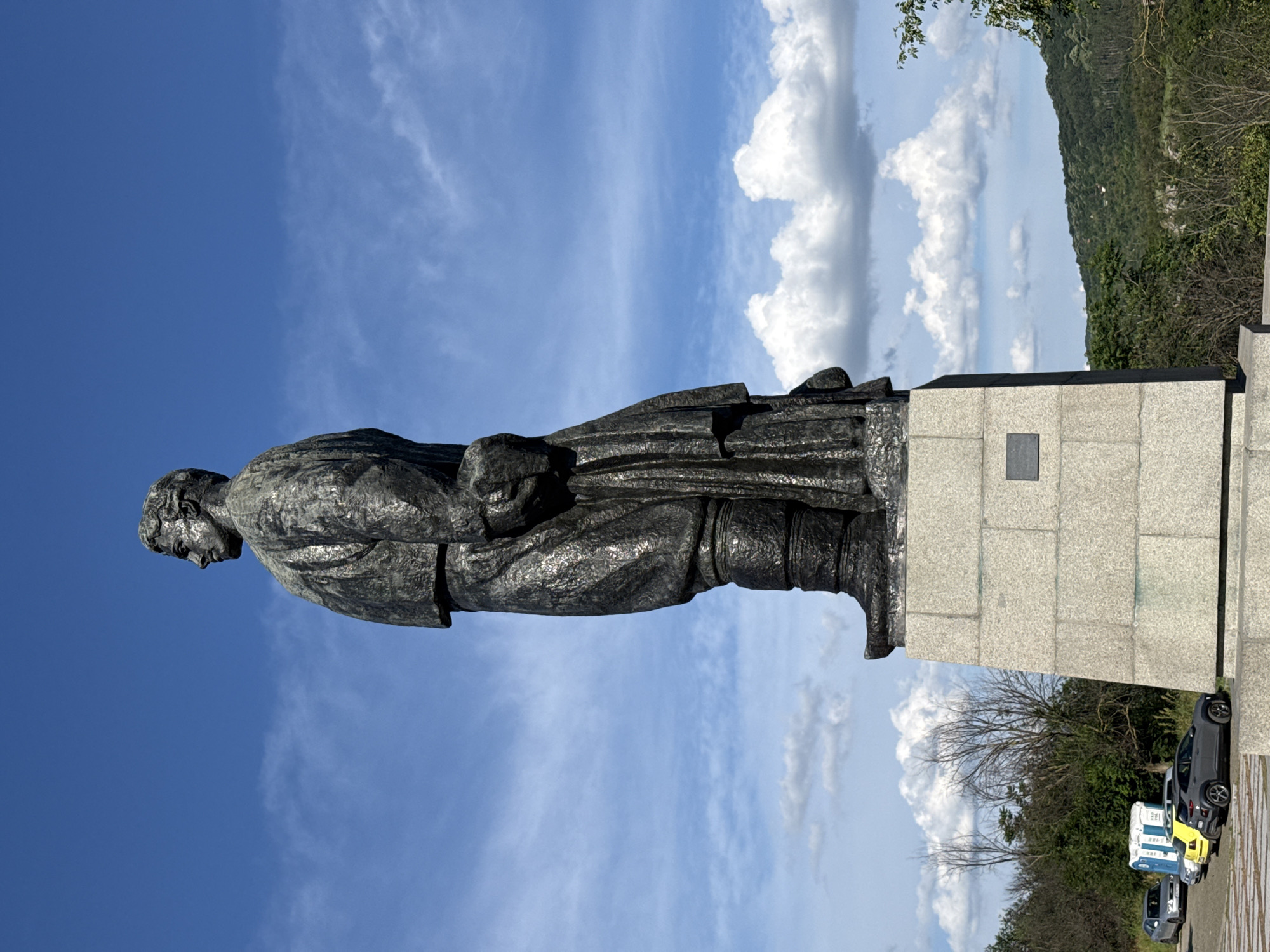 The image depicts a large, dark bronze statue of a standing man, viewed from the side and slightly from below. The statue is mounted on a tall, multi-tiered granite pedestal. The man in the statue is wearing historical clothing, including boots, trousers, and a jacket or coat with a draped fabric element. His head is turned to his left, and he appears to be looking into the distance. The sky behind the statue is a bright blue with scattered white clouds. In the background, there are green trees and a gently sloping, tree-covered hillside. At the base of the pedestal, several cars are parked, including a silver sedan, a yellow hatchback, and a grey SUV. Two portable toilets are also visible near the parked vehicles. The overall scene suggests an outdoor monument located in a park-like or scenic area. This statue is the monument to Todor Kirkov, located in the city of Lovech, Bulgaria.