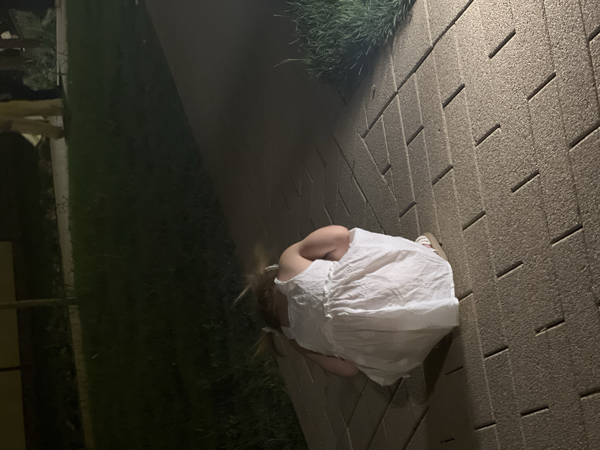 In the image, a young girl is seen from behind, wearing a white dress and crouching on a paved walkway. The walkway appears to be made of rectangular paving stones with grooves. To the left of the walkway is a patch of dark green grass, and beyond that, some indistinct structures and vegetation are visible, suggesting an outdoor setting. To the right of the walkway, there is a grassy area with tall blades of grass. The lighting in the image is dim, with artificial light illuminating the walkway and the girl, casting shadows. This indicates that it is likely nighttime or dusk. The girl is hunched over, with her knees drawn up and her head bowed, obscuring her face. Her blonde hair is visible, tied in pigtails. She is wearing light-colored sandals with stripes. The overall mood of the image is quiet and perhaps a little melancholic due to the girl's posture and the dim lighting. The setting appears to be a residential area or a park in Shymkent, Kazakhstan, given the provided location context. There is no visible text in the image.
