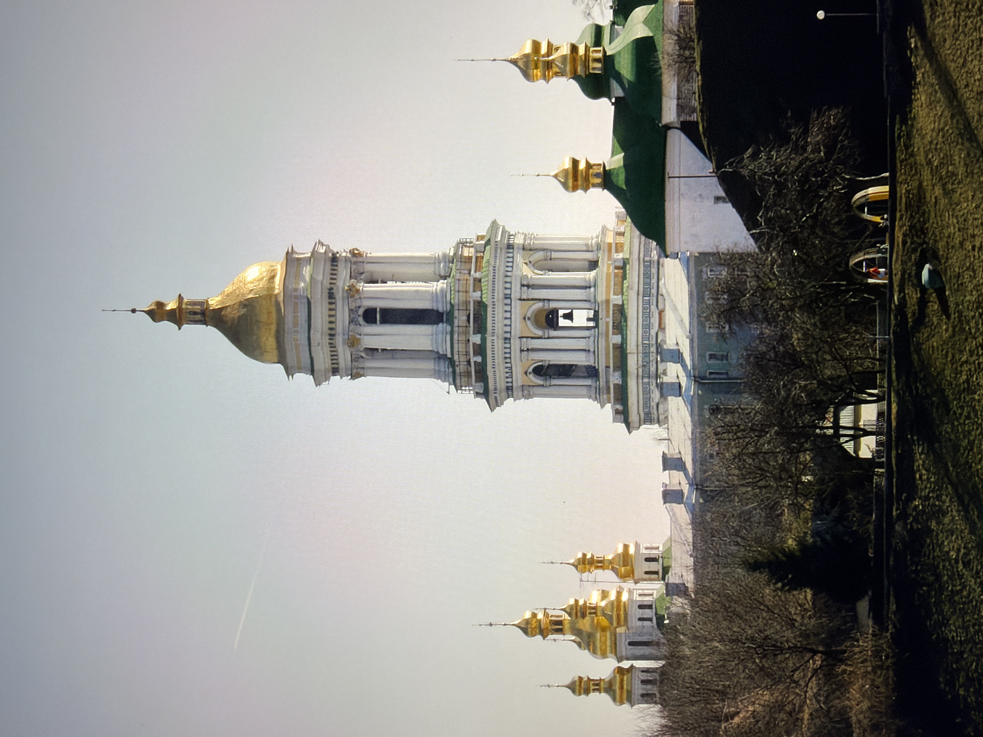 The image captures a panoramic view of the Kyiv Pechersk Lavra, a historic Orthodox Christian monastery in Kyiv, Ukraine. The main subject is the Lavra's bell tower, a tall, multi-tiered structure with a golden dome, topped with a cross. Surrounding the bell tower are several other smaller churches, also featuring golden domes. The buildings are predominantly white with ornate architectural details. The sky is a pale, hazy blue with a faint contrail visible from an airplane. In the foreground, there are leafless trees and a grassy slope. The overall scene suggests a bright, possibly spring or autumn day, with the monastery complex prominently displayed against the sky. The architecture is consistent with Eastern Orthodox churches, and the specific style and scale of the buildings are characteristic of the Kyiv Pechersk Lavra.