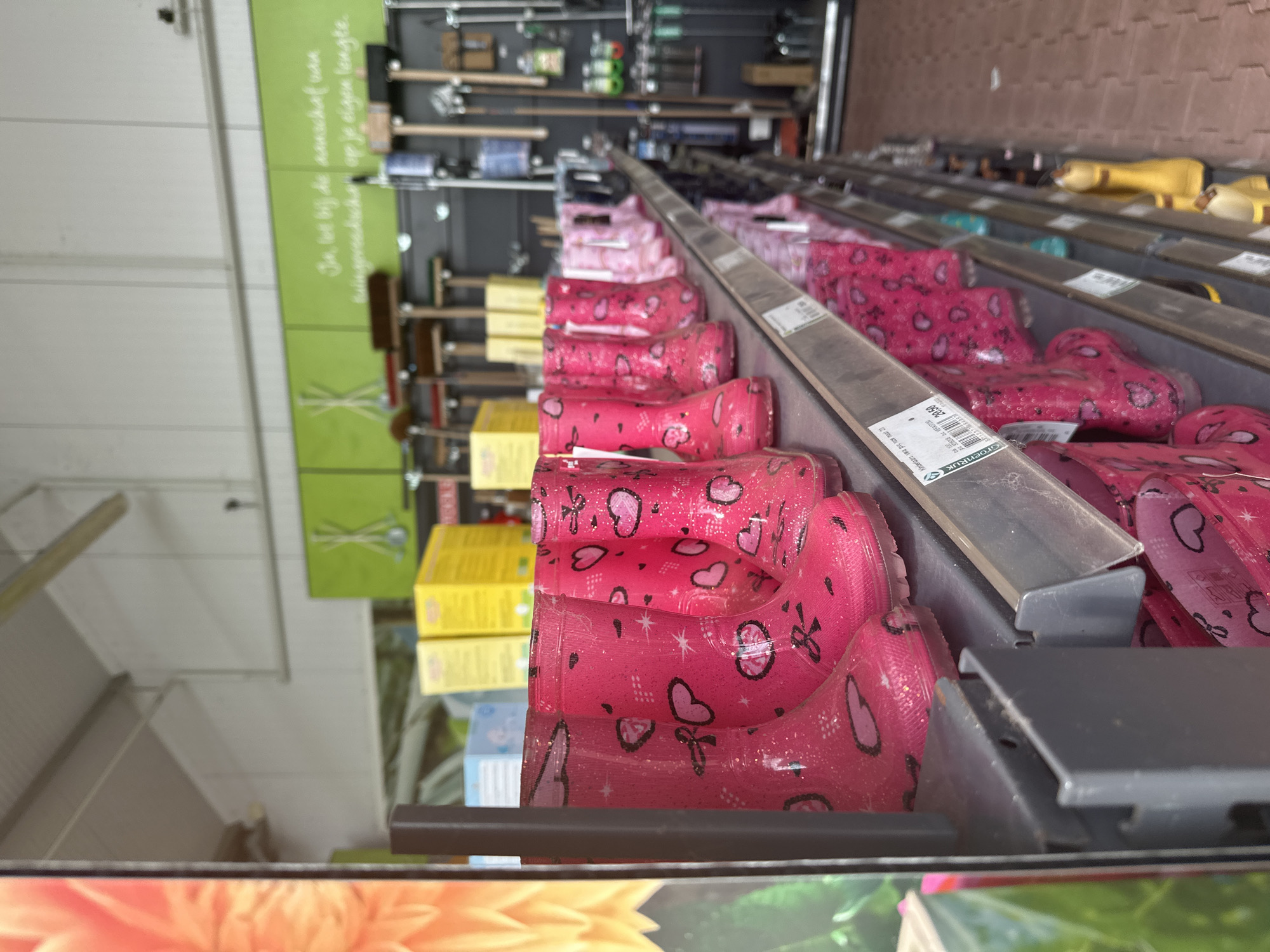 The image shows a retail display of pink children's rain boots with heart and star patterns, arranged on shelves in a store. The boots are stacked in rows, with the front row being the most prominent. Behind the boots, various gardening tools like rakes and brooms are visible on a dark wall. There are also yellow boxes stacked on a shelf. A price tag with the text "Kindermom. Weg PVS. Nu maar 28,50" is attached to one of the shelves. The store appears to be a garden center. The image was taken inside a store located in Tuk, Netherlands, which is evident from the text on the price tag referencing "Groenrijk," a Dutch garden center chain.
