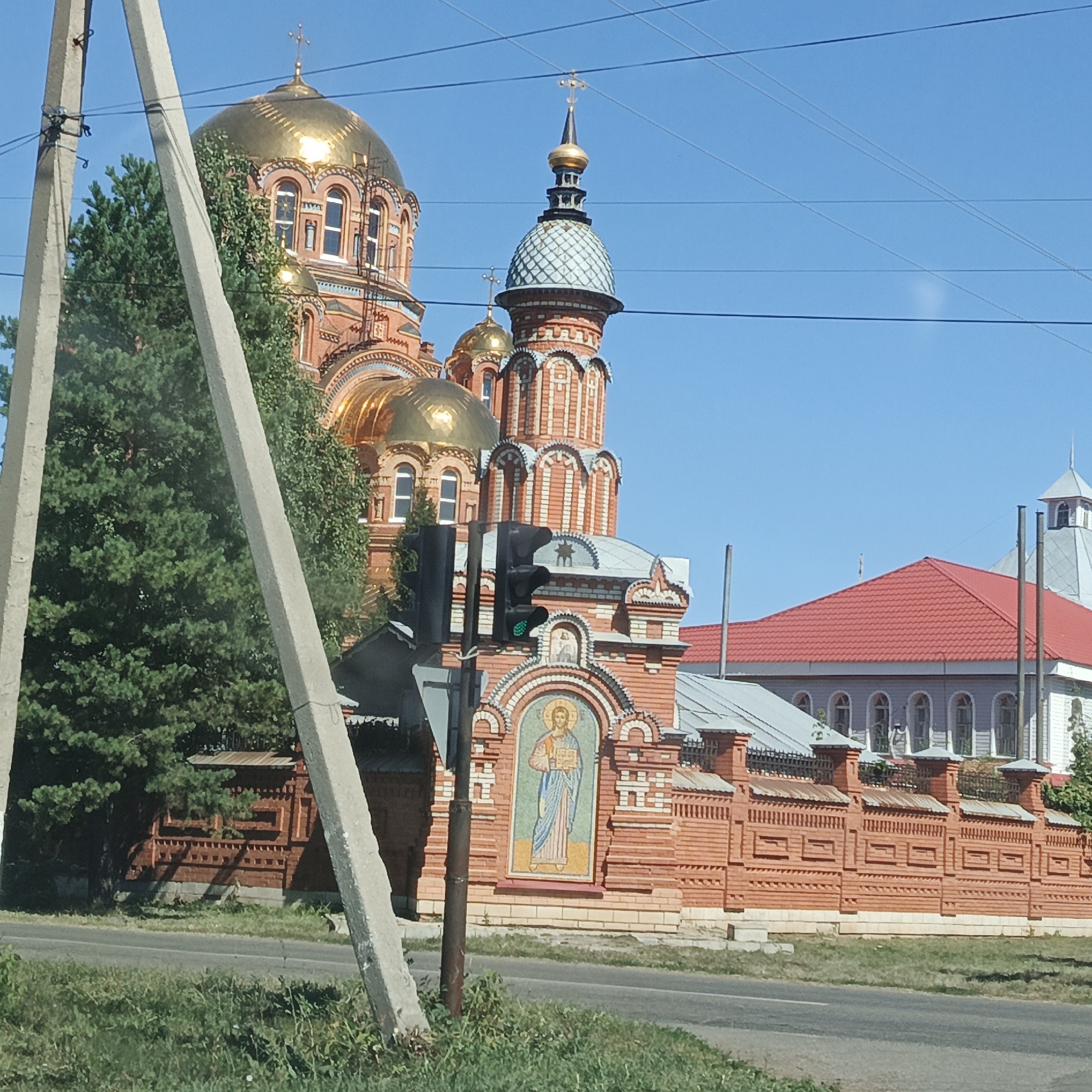 The image shows the Orthodox Church of the Transfiguration in Saraktash, Russia. The church is a large, red brick building with a prominent golden dome. There are also several other buildings visible, including a building with a red tiled roof and a building with white siding and arched windows. The church is surrounded by a red brick fence. The scene is captured on a sunny day with a clear blue sky. A traffic light is visible in the foreground, showing a green light. Two concrete utility poles stand in the left foreground. A road runs in front of the church, with grass and vegetation along the roadside.