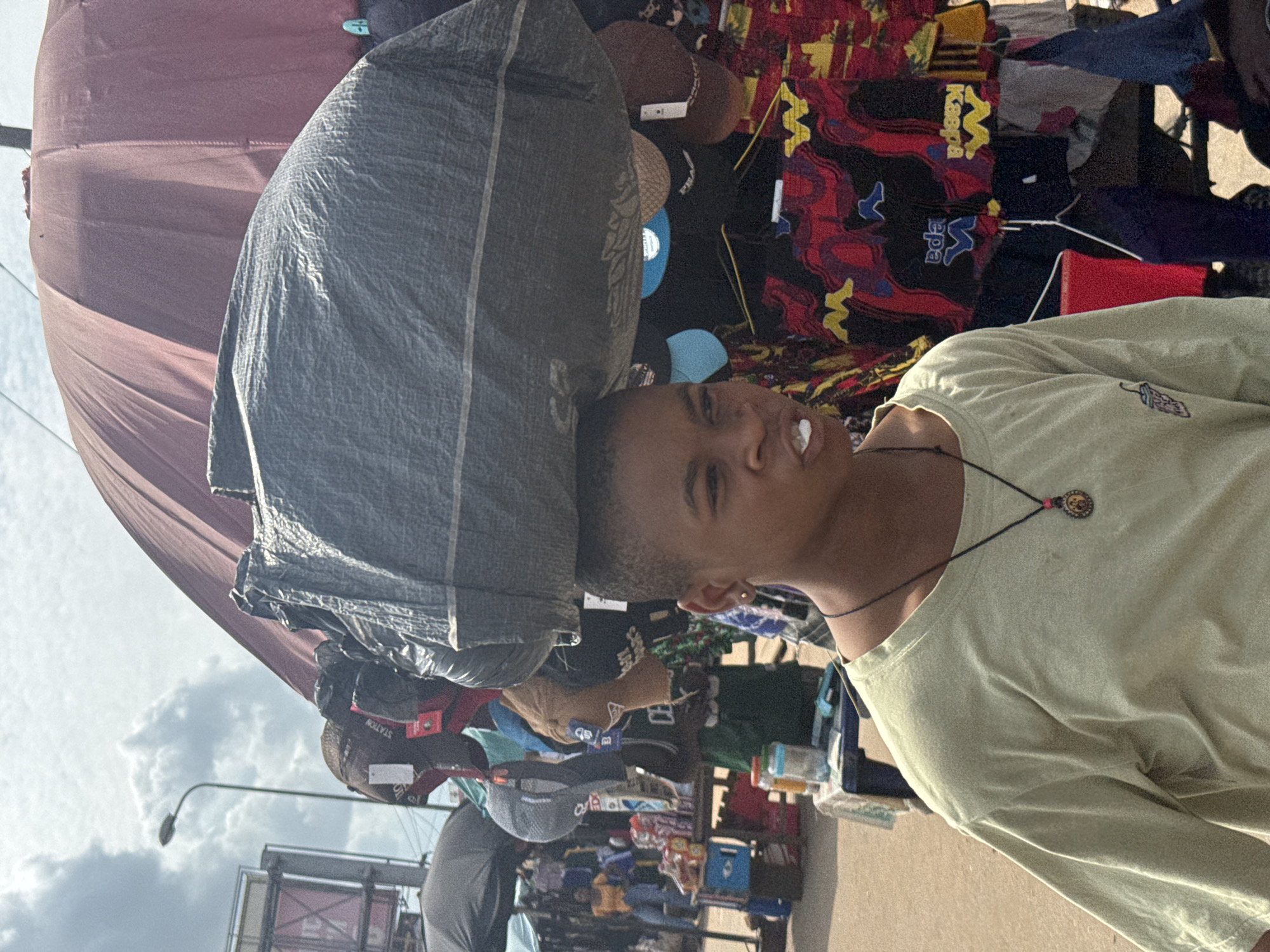 In Nkpor, Nigeria, a young person is depicted walking through a bustling market. They are carrying a large, black plastic-wrapped bundle balanced on their head. The bundle appears to be heavy and is draped over a large, reddish-brown umbrella. The person is wearing a light green t-shirt with a small embroidered image of a cup on the left chest and a dark pendant necklace with a circular charm. Their facial expression is neutral, with their mouth slightly open, and they have a single white object, possibly food, visible in their mouth.
The background reveals a vibrant market scene. Stalls are set up with various goods, including hats displayed on racks, colorful fabrics with abstract patterns, and bottled items on tables. Several people are visible in the background, engaged in market activities. A street lamp and a partial view of a billboard can be seen in the upper left of the image. The ground is a dusty, reddish-brown, typical of a dirt market. The overall atmosphere suggests a typical day in a busy Nigerian marketplace.
