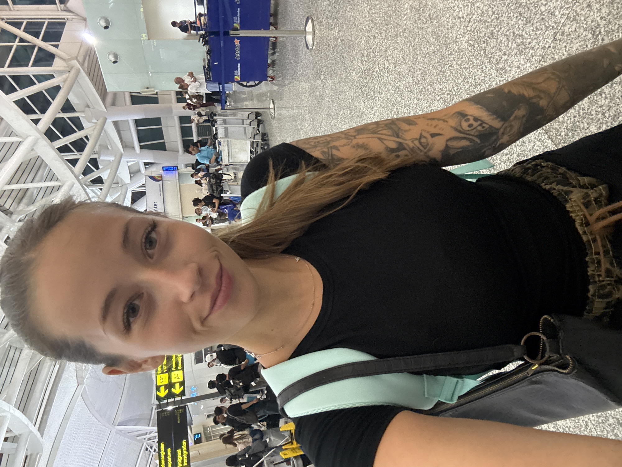A woman is taking a selfie at what appears to be an airport. The woman has fair skin, light brown hair, and is wearing a black shirt. She has a tattoo on her left arm. The airport has immigration signage, indicating the location is in Kuta, Indonesia. There are other people in the background, some wearing masks. There is a metal barricade and a Jetstar sign visible. The ceiling has a modern, geometric design.