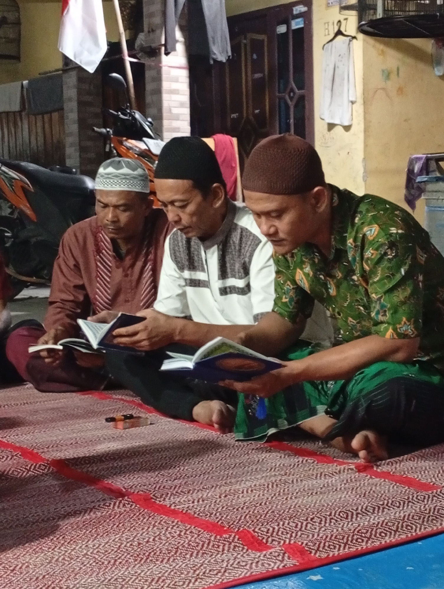 Three men are seated on a red and white woven mat. They are facing towards the left of the frame, with their heads bowed as they read from open books. The man on the far left wears a maroon robe and a white and grey patterned skullcap. He is holding a small, open book. The man in the center wears a white shirt with grey horizontal stripes and a black skullcap. He is holding a larger, open book. The man on the far right wears a green, patterned shirt with a brown skullcap. He is holding an open book. All three men appear to be focused on their reading. The background shows a building with yellow walls and a dark brown door. A motorcycle is visible to the left, and a flag, possibly the Indonesian flag, is hanging from a pole. The scene appears to be outdoors or in an open-air area.