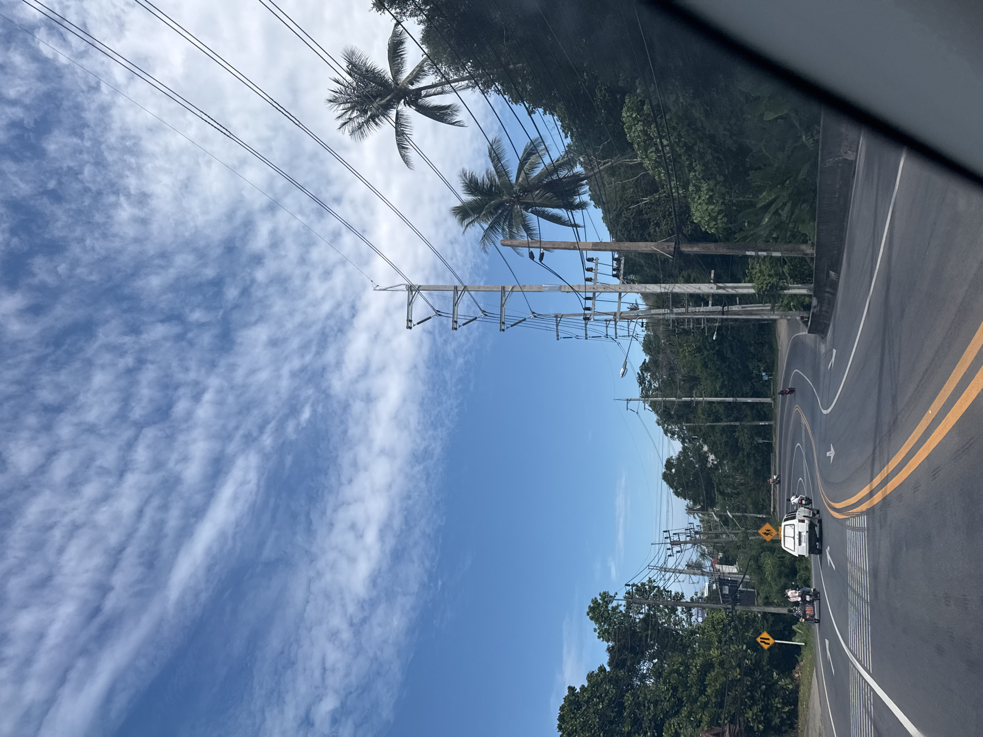 The image shows a curved road with traffic. In the foreground, a white van is driving in the right lane, followed by a motorcycle. To the left of the van, a motorcycle with a sidecar is moving in the left lane. The road is bordered by trees and vegetation on both sides, with power lines running overhead. The sky is blue with scattered white clouds. The overall scene appears to be in a tropical or subtropical location, given the presence of palm trees and lush greenery. The road has clear lane markings, including solid yellow lines and dashed white lines. There are yellow warning signs on the side of the road indicating potential hazards. The lighting suggests it is daytime, possibly late morning or early afternoon.