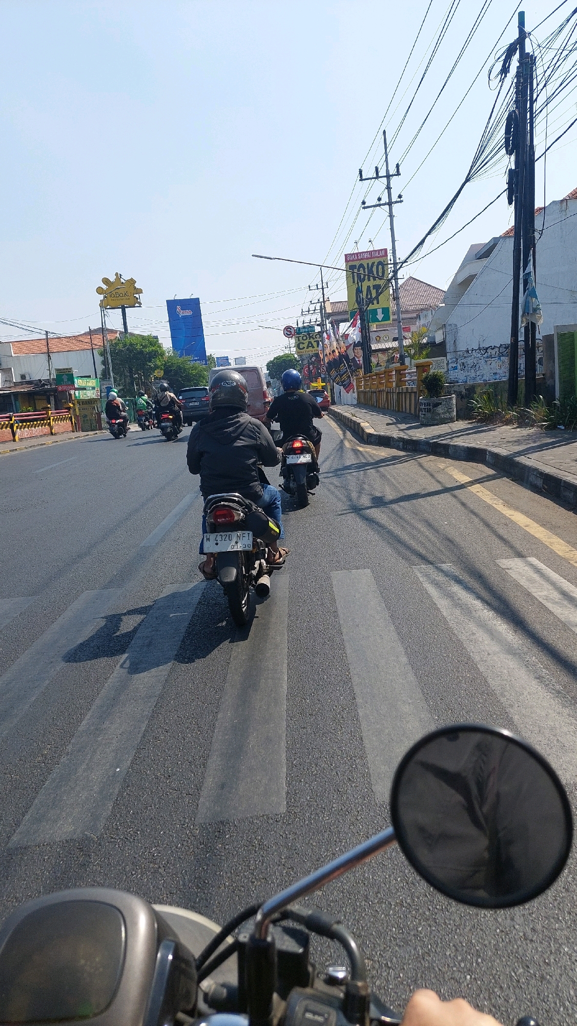 The image depicts a street scene in Sidoarjo, Indonesia, likely on a bright, sunny day given the clear sky and strong shadows. The perspective is from the rider of a motorcycle, as indicated by the handlebars and rearview mirror in the foreground.
Several motorcycles are traveling on a paved road with clear lane markings. One motorcycle, with the license plate W 4320 NFI, is directly in front of the viewer, with a rider wearing a black jacket and a black helmet. To the right and further ahead, another motorcycle rider with a blue helmet is visible. In the background, more motorcycles and some cars are moving along the road.
Along the right side of the road, there is a sidewalk with some vegetation. Buildings are visible behind the sidewalk, including one with a white facade and another with red tile roofs. Several signs are also present, including one with "TOKO GAT" and another with "BUKA SAMPAI MALAM". Utility poles with a dense network of wires are a prominent feature of the urban environment. On the left side of the road, a yellow building with a sign that appears to say "BAKERY" is visible. A pedestrian crossing is marked on the road, which the motorcycles are approaching or crossing. The overall impression is a typical urban street with moderate traffic.
