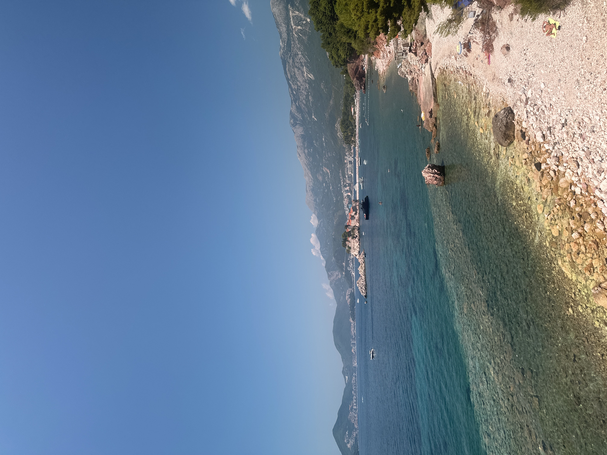 The image depicts a coastal scene in Sveti Stefan, Montenegro, on a clear, sunny day with a vibrant blue sky. The foreground features a pebble beach with scattered rocks and sparse green vegetation. A few people are visible on the beach, some sunbathing on towels, while others are near the water. The sea is a striking turquoise color near the shore, gradually deepening to a rich blue. The water is clear enough to see the rocky seabed in shallower areas.
In the middle ground, the rocky islet of Sveti Stefan is visible, connected to the mainland by a narrow causeway. The islet is densely packed with stone buildings and appears to be a historical landmark. Several boats, including a large dark yacht and smaller speedboats, are moored in the bay surrounding the islet and further out at sea. The coastline extends to the left, with a town or village nestled along the hills.
The background is dominated by a mountain range, lush with green trees, rising steeply behind the coastal settlements. The mountains are textured with visible rock formations and patches of lighter vegetation. The overall impression is a picturesque coastal resort area, combining natural beauty with historical architecture.