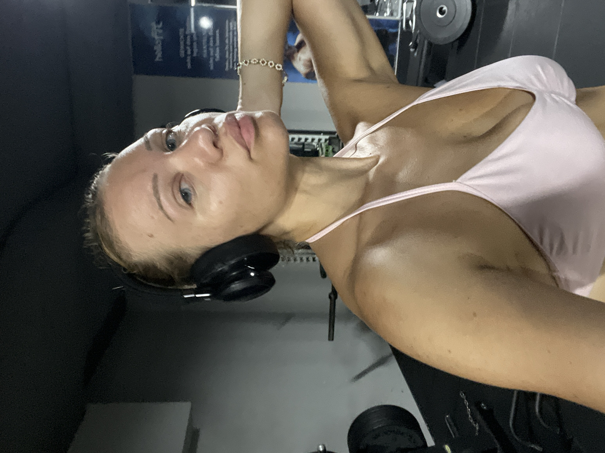 A woman is seen from the chest up, wearing headphones and a pink sports bra in what appears to be a gym. The background is dimly lit with gym equipment visible, including weights and a sign that reads "hellofit". The woman has light skin, blonde hair pulled back, and is looking slightly off-camera with her right arm behind her head. The image is a selfie taken in Mühldorf a. Inn, Germany.