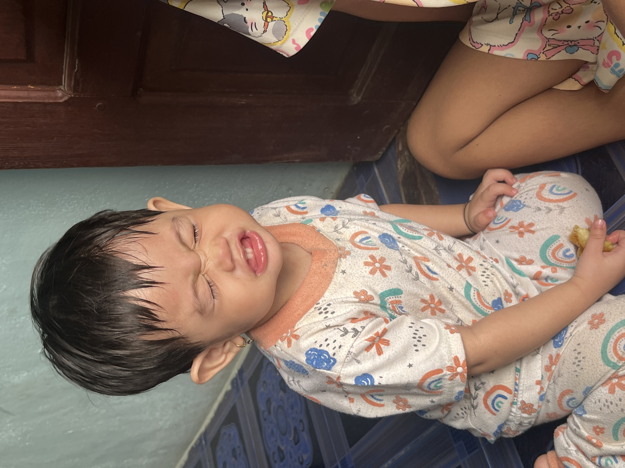 A young child, likely a toddler, sits on a tiled floor in what appears to be an indoor setting. The child has dark, wet hair and is wearing pajamas adorned with a pattern of rainbows and flowers. The child's eyes are squeezed shut, and their mouth is open, revealing their tongue and teeth, suggesting they are crying or making a distressed face. One of the child's ears is visible, with a small hoop earring. A piece of food, possibly a cracker or a biscuit, is held in the child's right hand. To the right of the child, another person's legs are visible, also wearing patterned clothing. The background includes a turquoise-colored wall on the left and a dark brown wooden door on the right. The floor is tiled with a blue and black pattern. The image is an indoor scene in Meliau Hulu, Indonesia, possibly a residential setting.