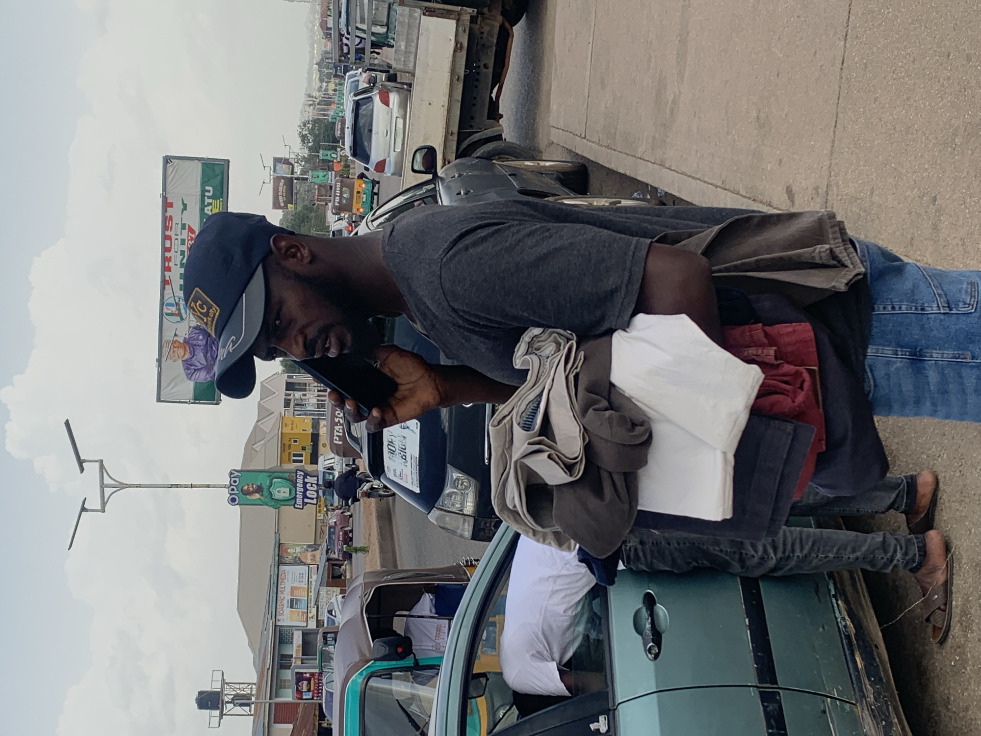 A Black man in a dark gray t-shirt and blue jeans is standing on the side of a street, holding a stack of folded clothes. He is also holding a black smartphone to his ear, appearing to be in conversation. He is wearing a dark blue baseball cap with a logo on the front. To his left, a teal colored car is parked, and a person in a white t-shirt is leaning out of the driver's side window. The man's feet are bare except for brown sandals. In the background, traffic is visible, including a light green auto-rickshaw, several cars, and a truck. Billboards with advertisements are posted on the side of the road. The environment appears to be a street in Gura Top, Nigeria, with a cloudy sky overhead.