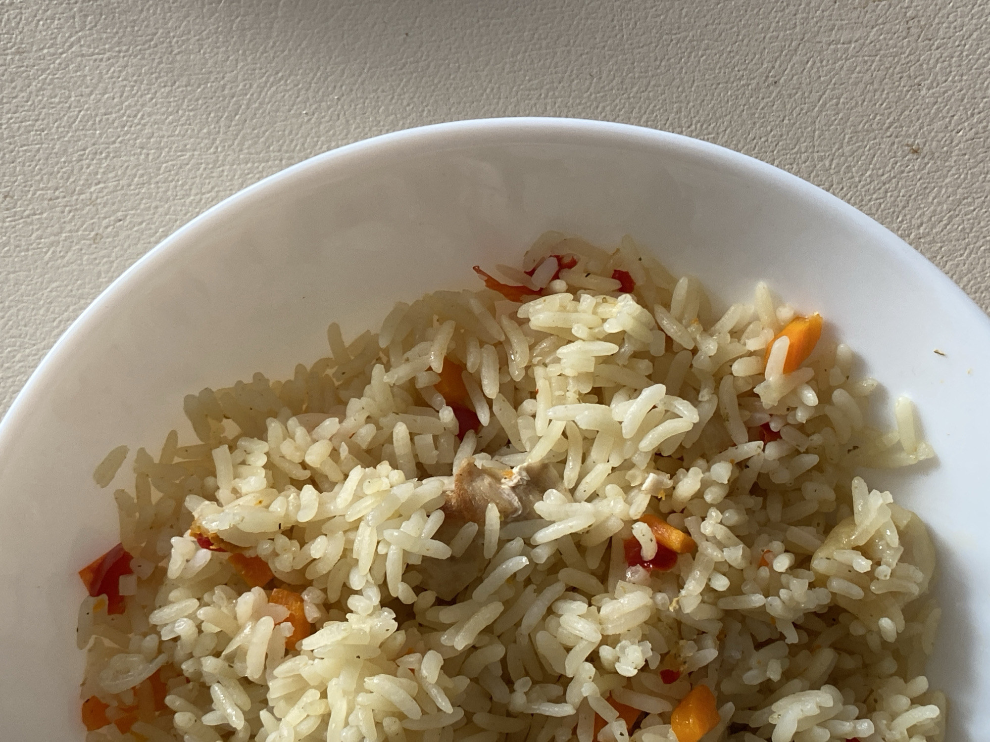 A close-up, overhead shot shows a white bowl filled with rice and diced vegetables, including carrots and possibly red bell peppers. The rice appears to be cooked and fluffy, mixed with small pieces of what might be chicken. The bowl is placed on a textured, light-colored surface, possibly a table. There is no indication of the city of Ostrovki, Russia.
