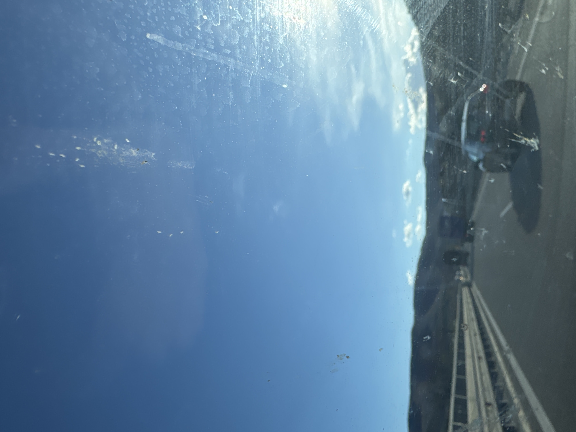 The image shows a view through a dirty car window. Outside the window is a bright blue sky with some clouds. On the right side of the frame, there is a blurred view of a highway, possibly an elevated section or bridge, with a car traveling in the same direction. The location may be near Dolno Varshilo, Bulgaria, based on the user's prompt. The window itself is covered in spots and smudges, which detract from the clarity of the outside scene.