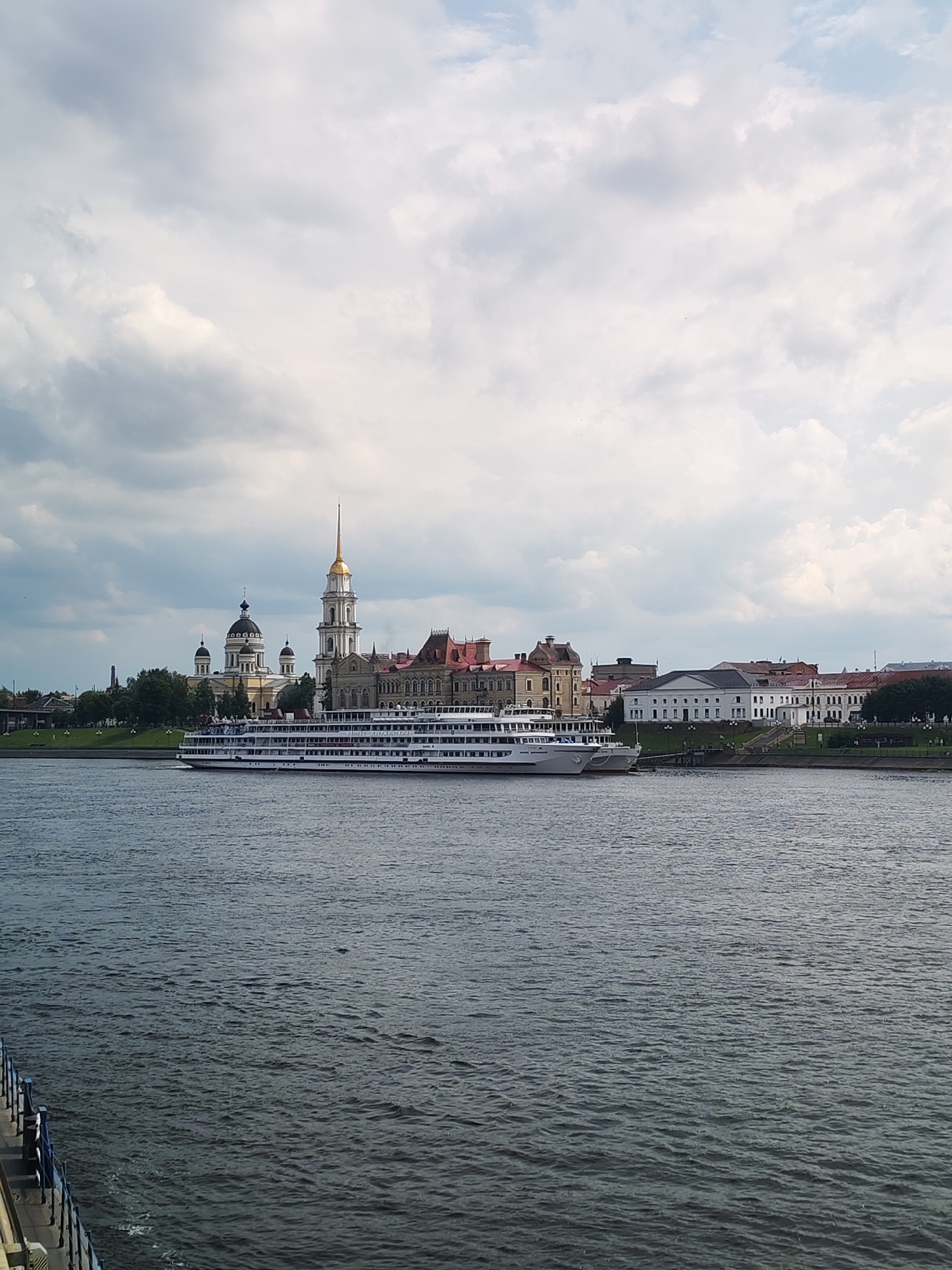 A wide, dark blue river occupies the foreground, with small ripples on its surface. In the middle ground, a large white river cruise ship is docked. Behind the ship, a complex of buildings with red tile roofs and white walls lines the riverbank. A prominent tall white tower with a golden spire rises above the buildings, and several other dome-shaped structures are visible, suggesting a religious complex. Green trees are scattered behind the buildings. The background is a cloudy sky with patches of blue. A small portion of a blue railing from another vessel is visible in the lower left corner. The scene depicts a cityscape along a river in Bol'shie Mkhi, Russia, under a partly cloudy sky. There are no people clearly visible in the image.
