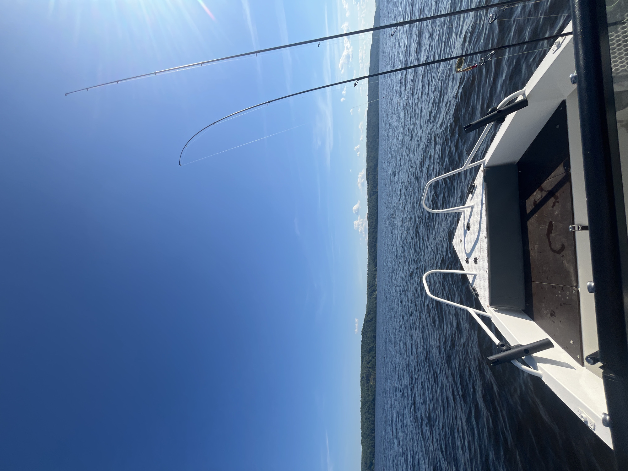 The image shows the bow of a white motorboat on a dark blue body of water. Two fishing rods are propped up on the right side of the boat, with fishing lines extending into the water. The sky is a bright, clear blue with a few wispy clouds and the sun is shining, casting a glare on the right side of the image. In the distance, a tree-covered shoreline stretches across the horizon. The location is identified as Siukterka, Russia.