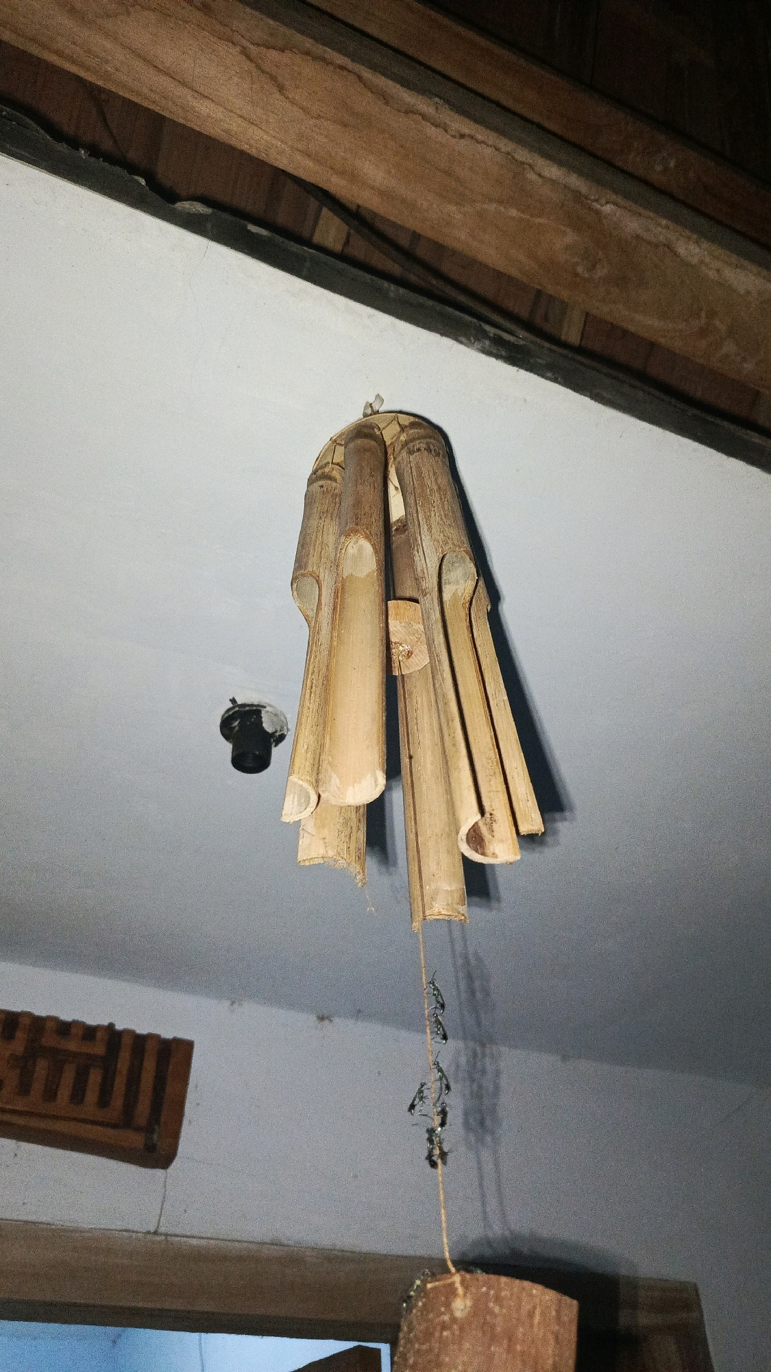 A bamboo wind chime hangs from a wooden beam on the ceiling of a room in Babadan, Indonesia. The wind chime is made of several hollow bamboo tubes of varying lengths, tied together at the top. A thin string dangles from the center of the wind chime, holding a small, dark object at the end. To the left of the wind chime, a black electrical socket is visible on the white ceiling. A wooden carving with intricate geometric patterns is mounted on the wall below the ceiling beam, to the left of the frame. The room appears to be under renovation or under construction, with exposed wooden beams and a plain white ceiling.
