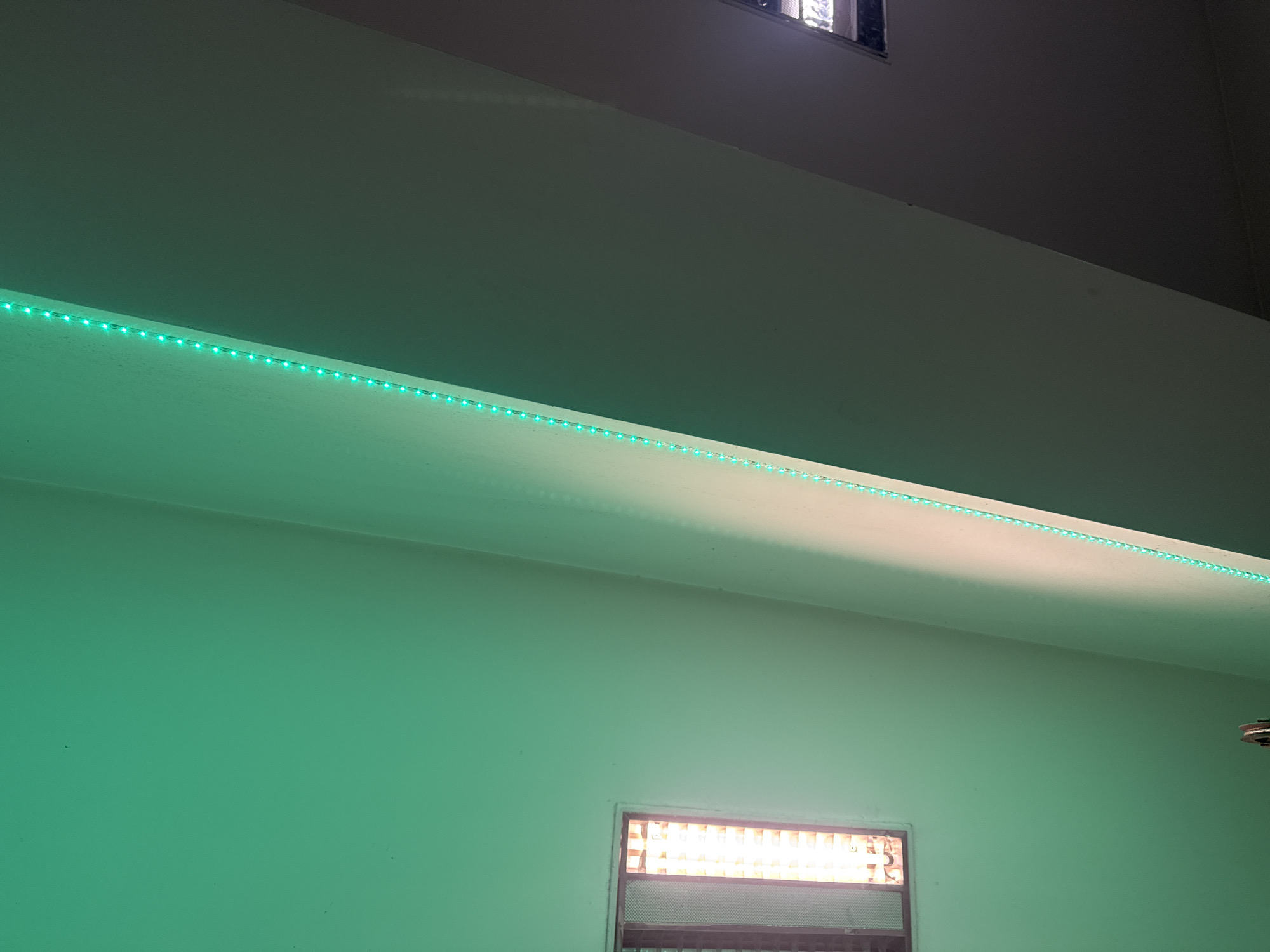 The image shows an interior view of a room. A strip of green LED lights runs along the edge of a ceiling ledge. Below the ledge, the wall appears to be painted in a light green color. There is a window near the bottom of the frame, emitting a bright orange glow from within. Above the ledge, the ceiling is a darker shade of gray or white. The overall lighting conditions suggest it is either nighttime or a dimly lit space. The location of the image is unknown, but given the request, the city is Varna, Bulgaria.