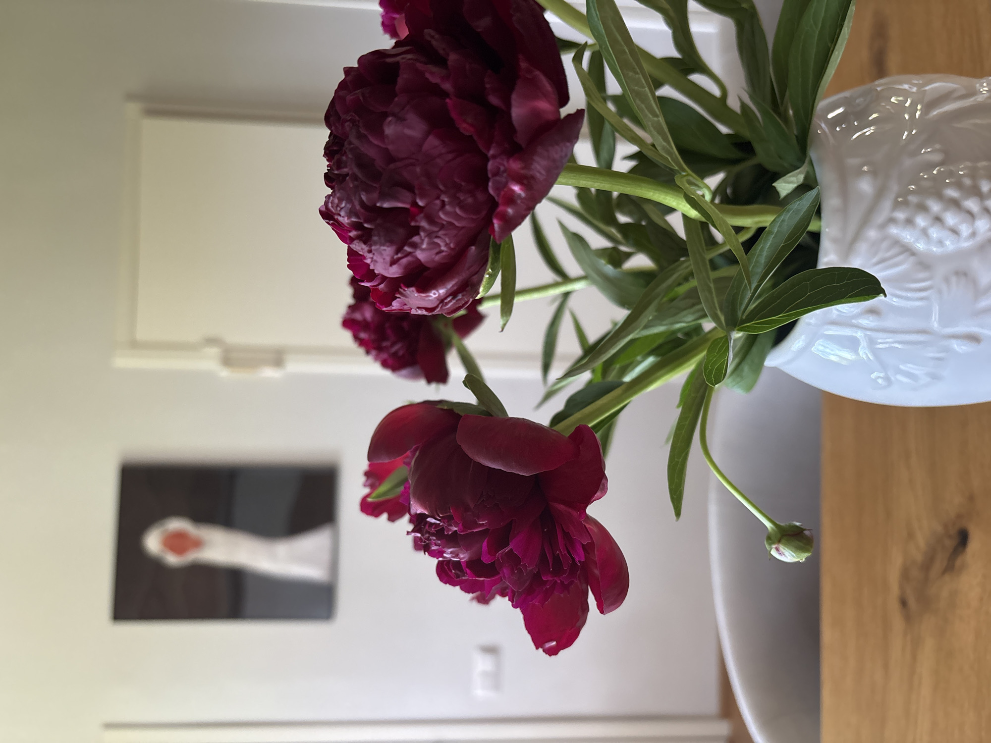 In the foreground, a white vase with a textured pattern, possibly depicting grapes, holds a bouquet of deep red peonies. The flowers are in various stages of bloom, with some fully open and others in bud. The vase sits on a wooden table. In the background, a white wall is visible, with a framed artwork hanging on it. The artwork is abstract, featuring a blurry image of a white swan-like figure with an orange beak against a dark background. To the right of the artwork, a white door frame is visible. The overall scene suggests an indoor setting, likely a home or a studio, bathed in soft natural light. No people are present in the image. There is no visible indication of the city of Chaikovsky, Russia, in the image.