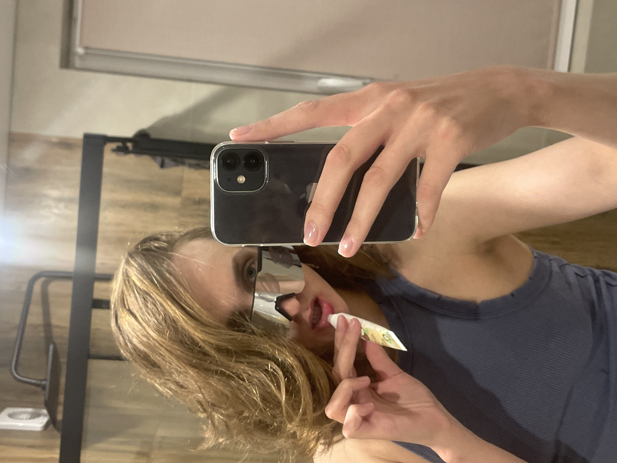 A person is taking a selfie in front of a mirror. The person is holding a black smartphone up in their right hand, with their left hand holding a small tube of lip balm. The person is wearing sunglasses. The location is Unknown, Unknown. The background shows a bathroom setting with a wooden floor.