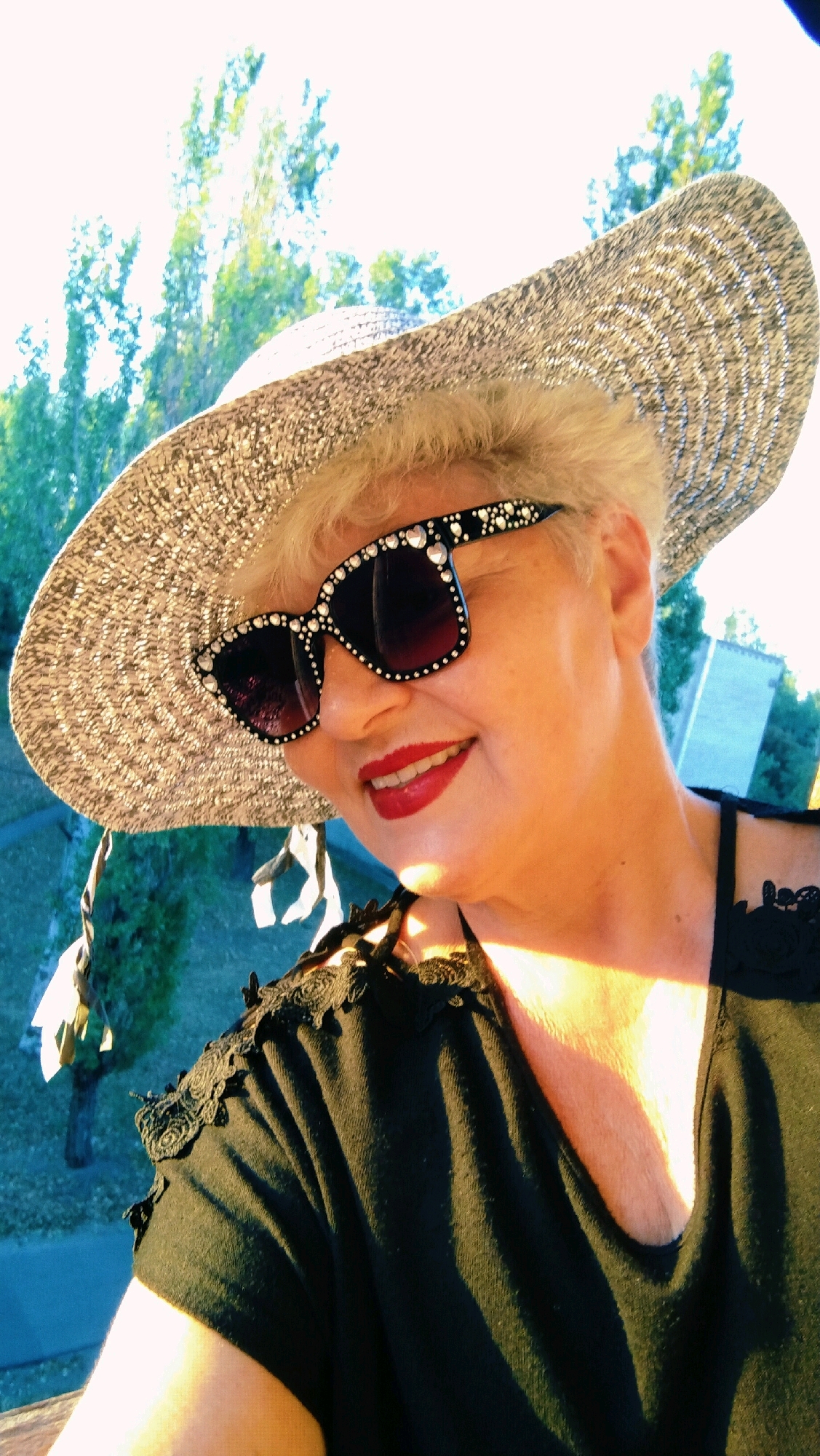 A medium shot shows a Caucasian woman with short blonde hair wearing a wide-brimmed straw hat and ornate sunglasses. She is smiling and looking slightly to her right. The woman is wearing a black top with lace trim on the shoulders. The background shows green trees against a bright sky, suggesting an outdoor daytime setting. There are no discernible landmarks to pinpoint the location in Horishni Plavni, Ukraine.