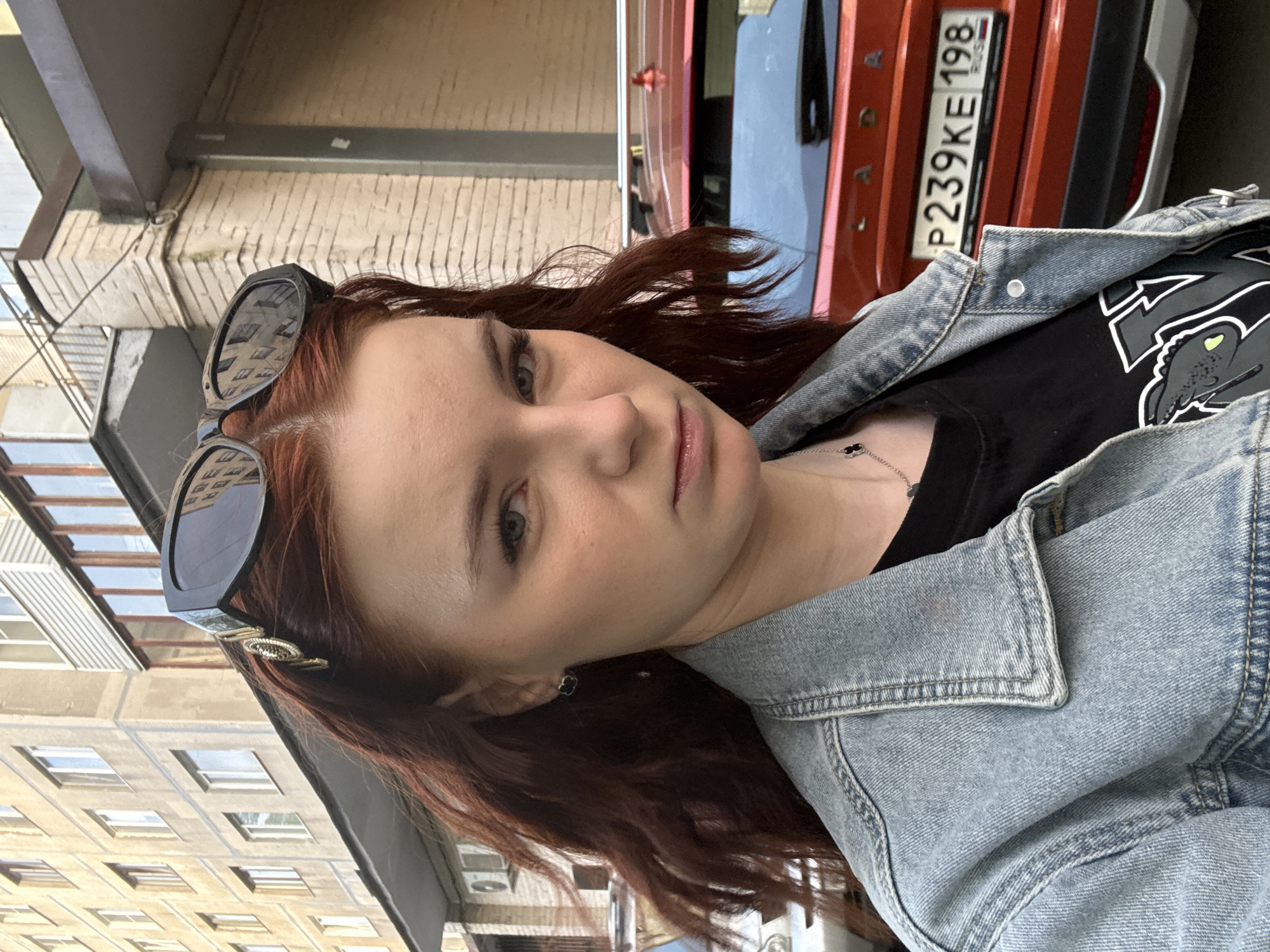 A woman with reddish-brown hair is posing outdoors. She has sunglasses pushed up on her head and is wearing a denim jacket over a black t-shirt with a graphic on it. She is wearing stud earrings. A vehicle, partially visible, has a license plate that reads "P239KE 198 RUS." The background shows a building constructed of light-colored brick. Based on the license plate information (198 RUS), this image was taken in Vsevolozhsk, Russia.