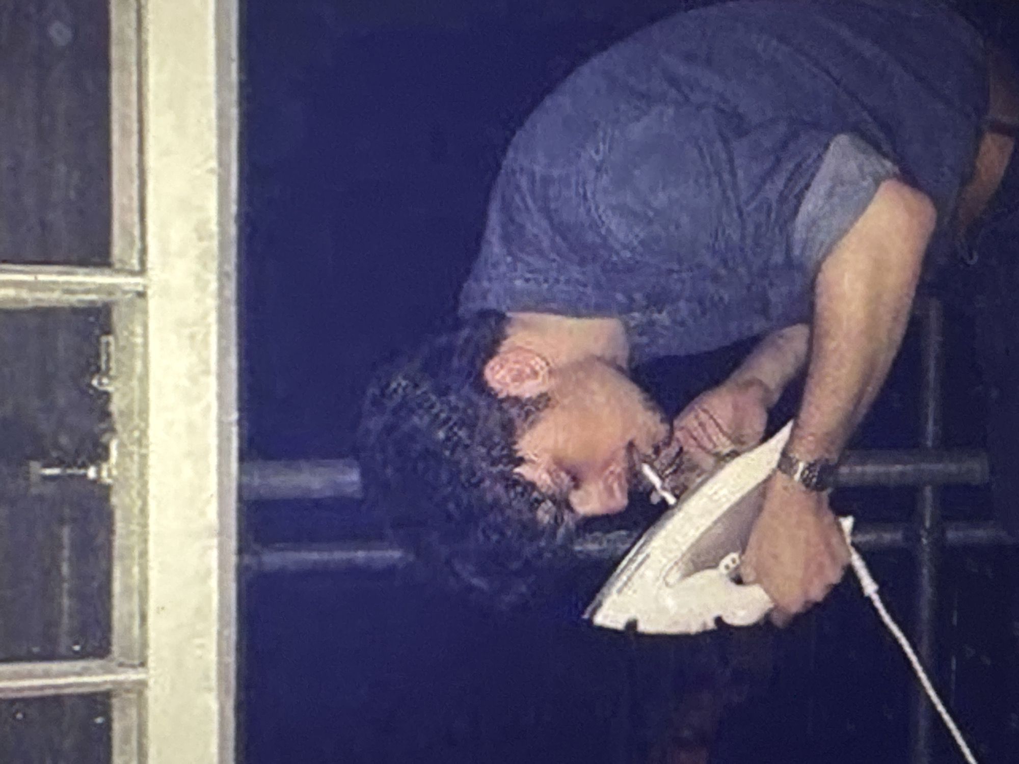 The media file depicts a person, likely male due to short hair and musculature, in a dimly lit environment. The individual is bent over, with their head down and eyes focused. They are wearing a dark blue, possibly denim, shirt and a watch on their left wrist.
In their hands, the person is holding a white iron. A lit cigarette is held between the fingers of their right hand, positioned near the iron. The posture and the presence of the iron and cigarette suggest an unusual or possibly dangerous activity, though the exact purpose is unclear from the image alone. The background is dark, with some blurry elements that could be railings or structures, hinting at an outdoor or industrial setting. The image is slightly grainy, typical of older digital or film photography, and the lighting suggests it was taken at night or in a poorly lit indoor space.
There are no visible text elements in the image. The overall mood is somewhat tense or secretive due to the dim lighting and the peculiar combination of objects and the person's posture. The location context provided, Kuta Selatan, Indonesia, does not offer specific visual cues within the image itself to confirm or deny this location.