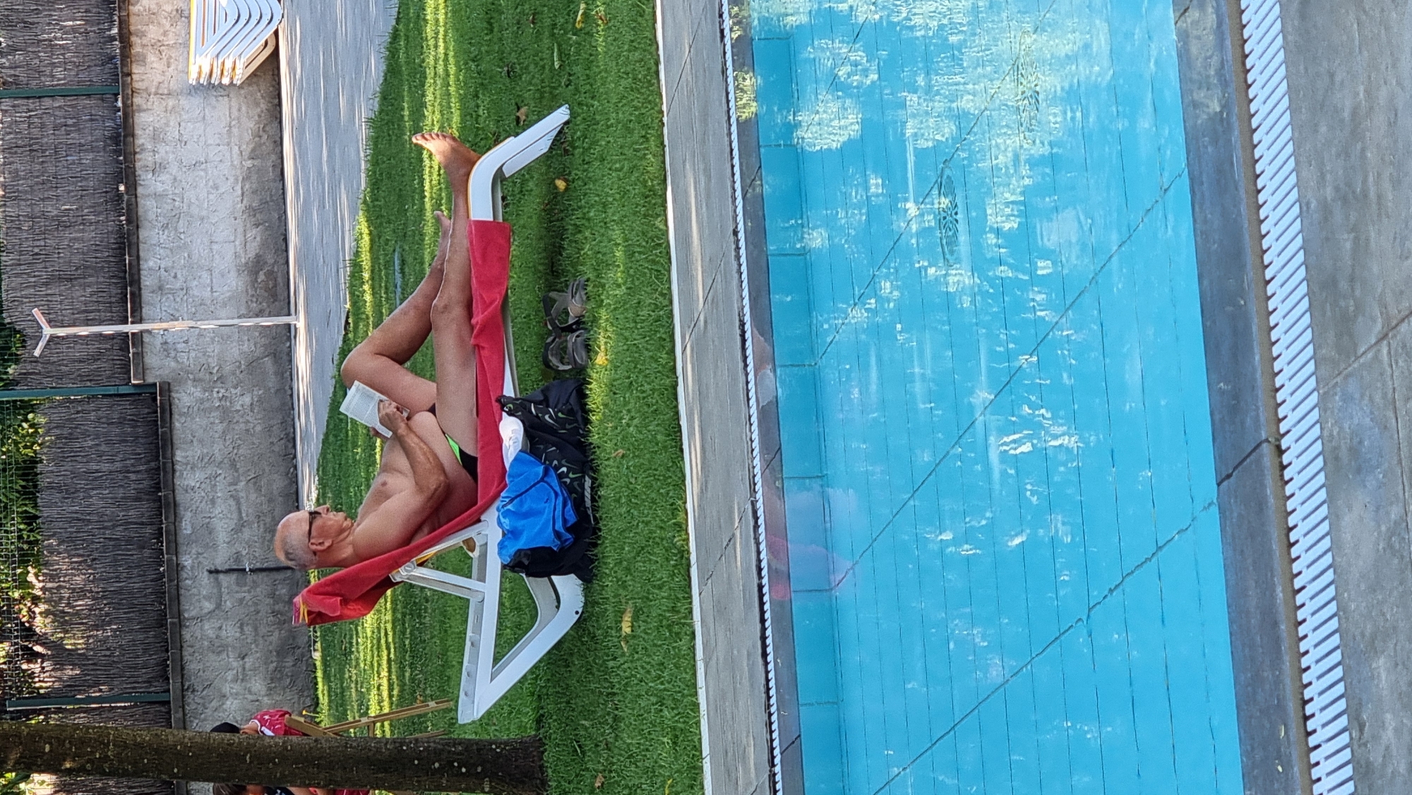 An older man with glasses and a shaved head is lying on a red towel on a white lounge chair by a swimming pool. He is wearing green swimming trunks and reading a book. A black bag, a blue shirt, and a pair of sandals are on the grass next to the lounge chair. The scene appears to be a leisure area, possibly a hotel or resort, in Sant Joan Les Fonts, Spain. The swimming pool water is blue and reflects the sky and surrounding environment.