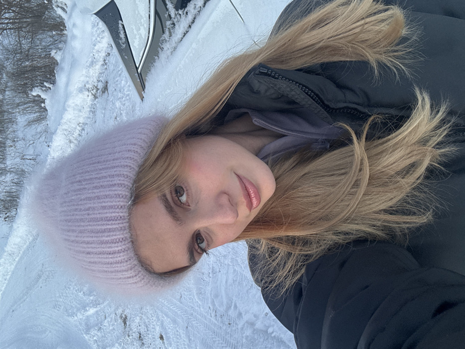This image displays a female individual in a snowy outdoor setting, located in the city of Unknown, Unknown. The subject, a young adult with light brown hair, is wearing a light purple fluffy knit beanie and a dark grey puffer jacket over a lighter grey or purple collared layer. She is depicted from the chest up, looking directly at the camera with a neutral expression and slightly parted lips. The background features snow-covered ground with visible tire tracks or indentations. To the subject's right, the top and side of a dark-colored vehicle, partially covered in snow, is present. Further in the background, a line of snow-covered bushes and bare trees is discernible.