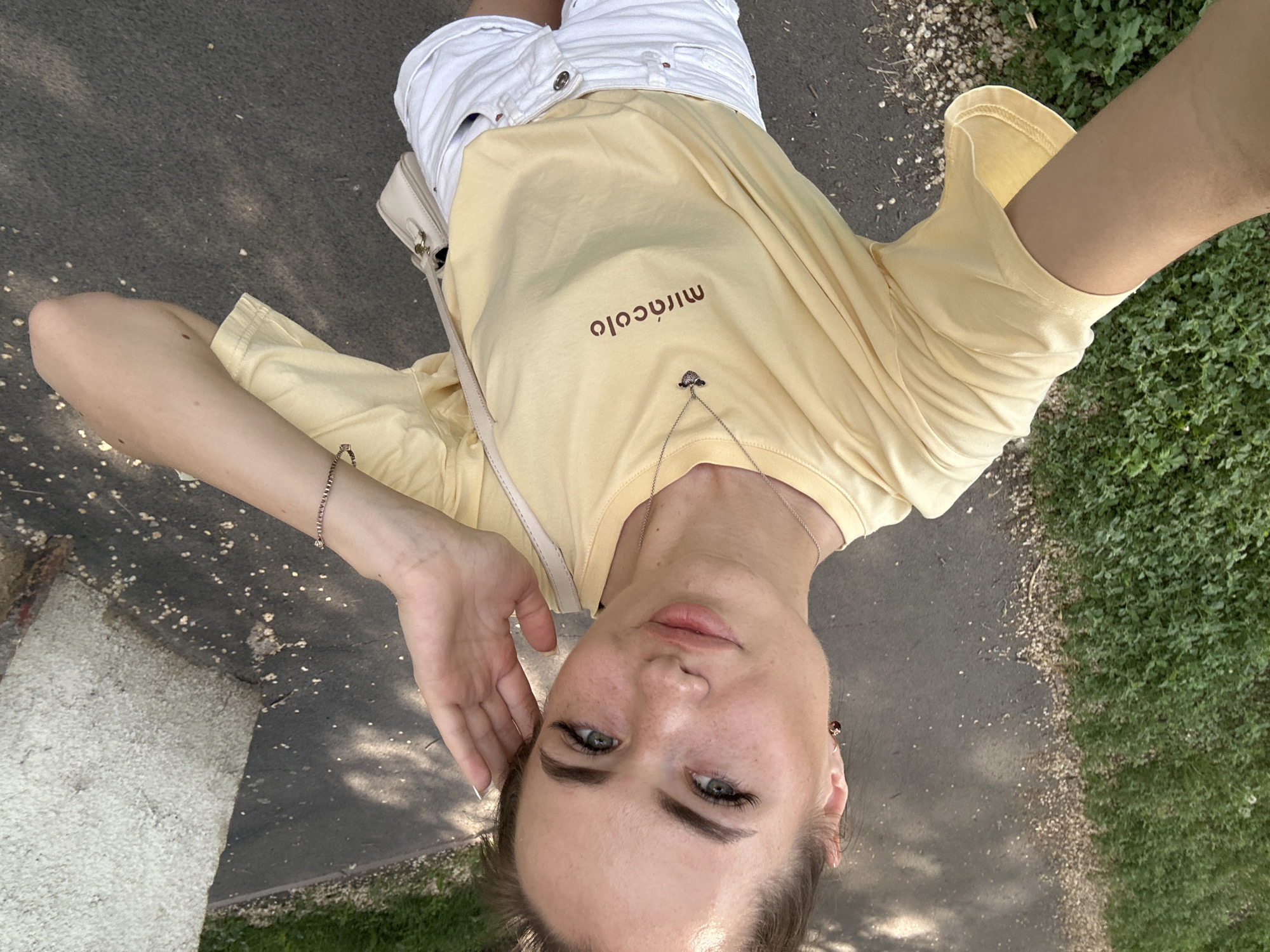 A woman is taking a selfie outdoors. She is wearing a light yellow t-shirt with the word "miracolo" printed in brown on the front and white denim shorts. A small beige purse is slung over her shoulder. She is holding her right hand to her ear and looking at the camera with a slight smile. The background shows a paved walkway and green grass. There is a concrete pillar to the right of the frame. The lighting suggests it is daytime.