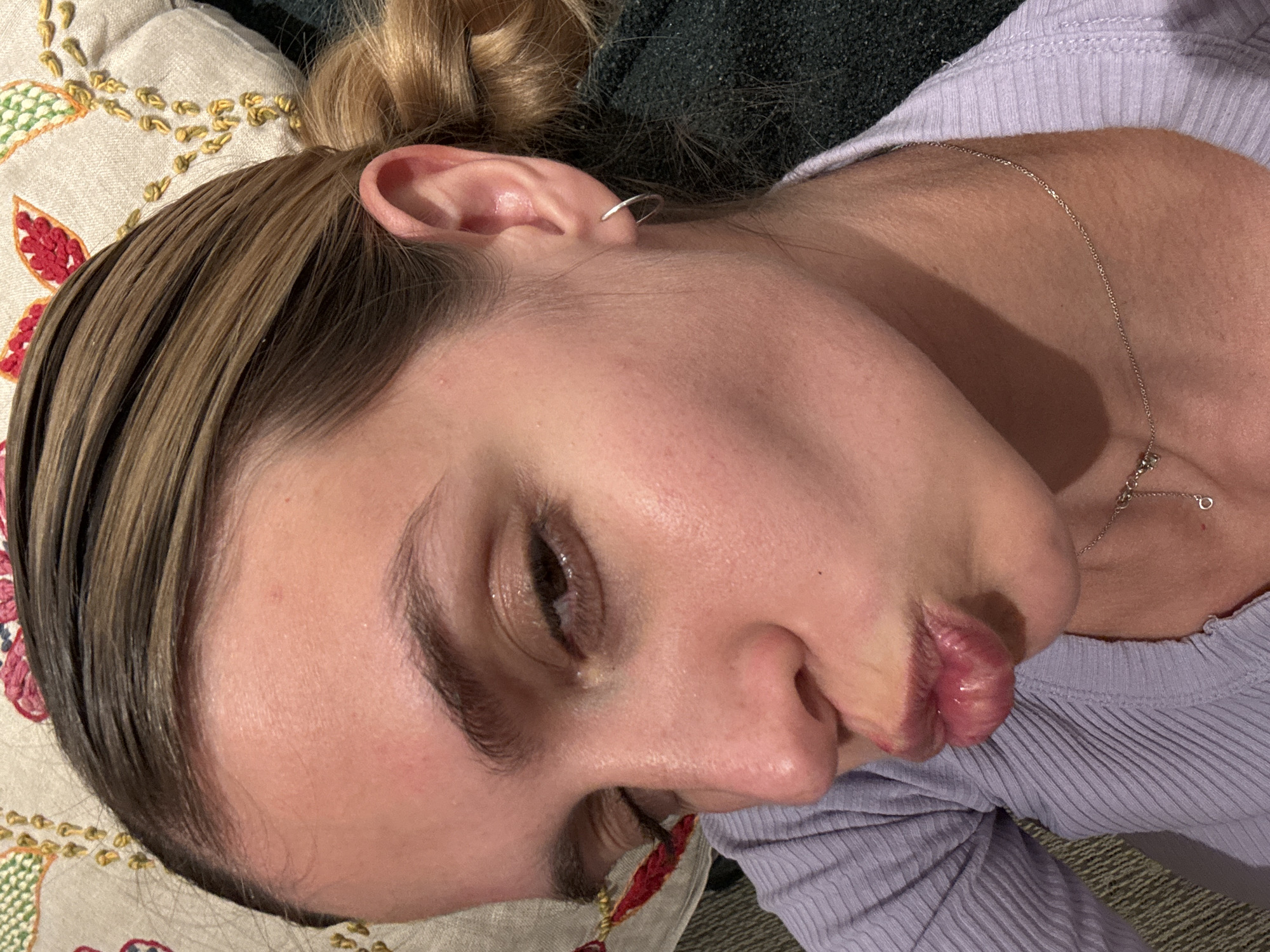 A close-up, angled shot captures a person, likely a young woman, lying down and looking towards the viewer. Her head is tilted, and her lips are puckered into a "fish-lips" expression, suggesting a playful or pouty mood. Her brown hair, with some lighter highlights, appears sleek or damp and is pulled back into a bun, visible at the top of her head.
She has clear skin and her eye shows minimal makeup. A small silver hoop earring adorns the helix of her left ear, and a delicate silver chain necklace with a small circular pendant hangs around her neck, resting on her collarbone. She is wearing a light purple, ribbed top.
The person is resting on a light-colored pillow embellished with embroidered floral patterns in red and yellow/gold threads. In the background behind her head, a dark, textured fabric, possibly a blanket or another piece of clothing, is visible. The lighting suggests an indoor setting, possibly with artificial light. There is no visible text in the image.