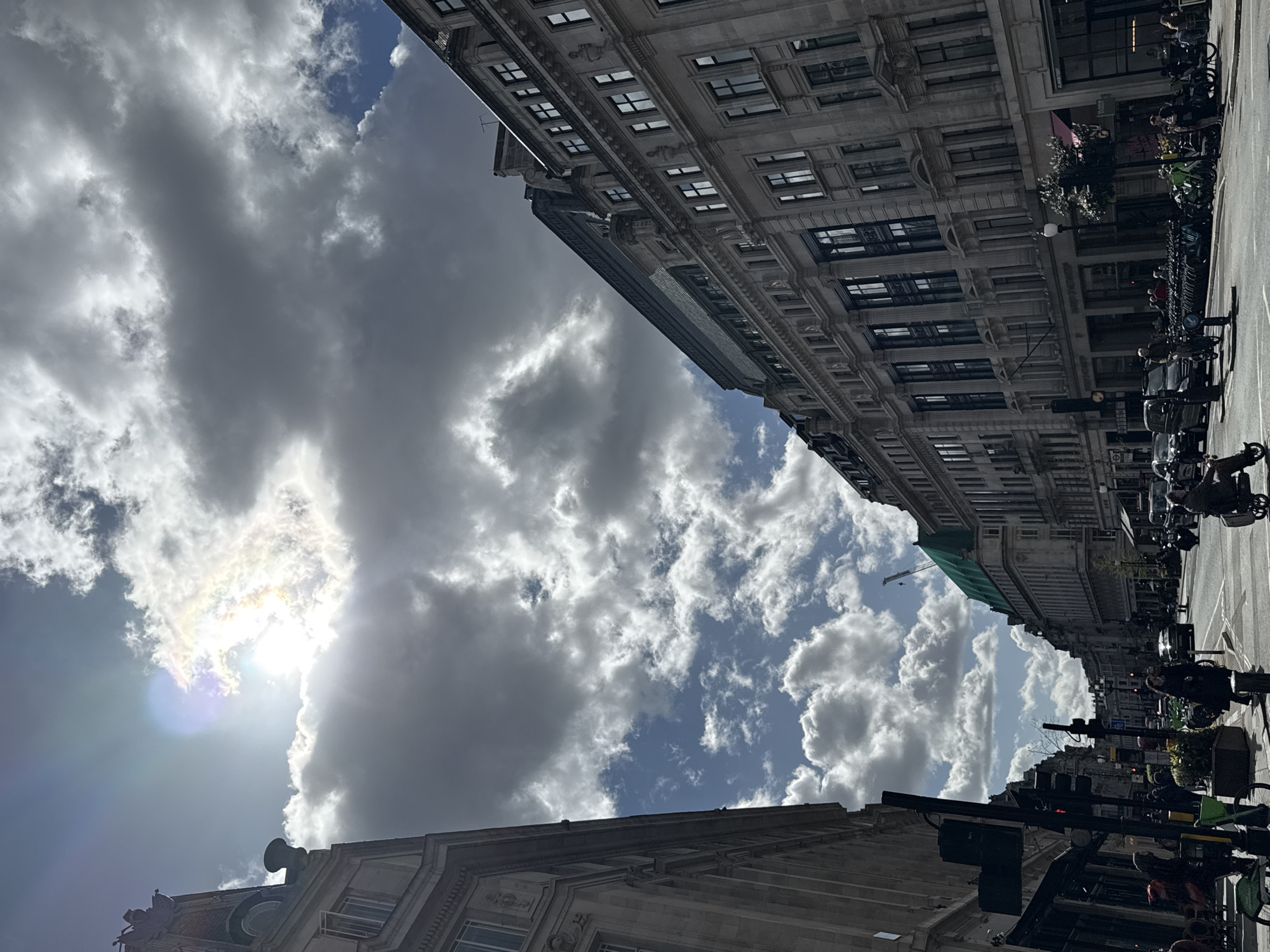 This image depicts a wide urban street in Knightsbridge, London, United Kingdom, captured during daytime under a dramatic, partly cloudy sky. The perspective is a low-angle shot, looking down a street flanked by multi-story, classically styled stone buildings.
The sky occupies the upper half of the frame, dominated by large, textured grey and white cumulus clouds. The sun is breaking through a gap in the clouds in the upper left quadrant, creating a strong lens flare and illuminating the cloud edges with bright white and subtle iridescence. Patches of deep blue sky are visible between the cloud masses.
On the left, a portion of a multi-story stone building with ornate architectural details, including statuary or decorative elements near the roofline and arched windows, is visible. Its facade is partially in shadow.
On the right, a prominent, long multi-story classical stone building extends along the street. This building features numerous regularly spaced rectangular windows, decorative pilasters or columns between sections, and a mansard-style roof. The name "HARVEY NICHOLS" is visible on its lower facade. Further down the street on the right, another building's upper section is covered by a large green construction tarp, with a tall crane visible behind it.
The street below is an asphalt road with visible lane markings. Vehicular traffic includes several cars (a black minivan and a black sedan) and motorcycles/scooters. In the immediate foreground, a person on a dark motorized scooter or moped is seen from behind, riding away from the viewer. A person on a bicycle is also visible further down the road. Traffic lights are present at intersections, with a red light visible in the distance.
Both sides of the street feature pavements populated by numerous pedestrians. On the right sidewalk, rows of parked bicycles and scooters are visible. Street furniture such as dark traffic signal poles and green planters with foliage are also present. On the left, ground-floor businesses with outdoor seating areas are visible.