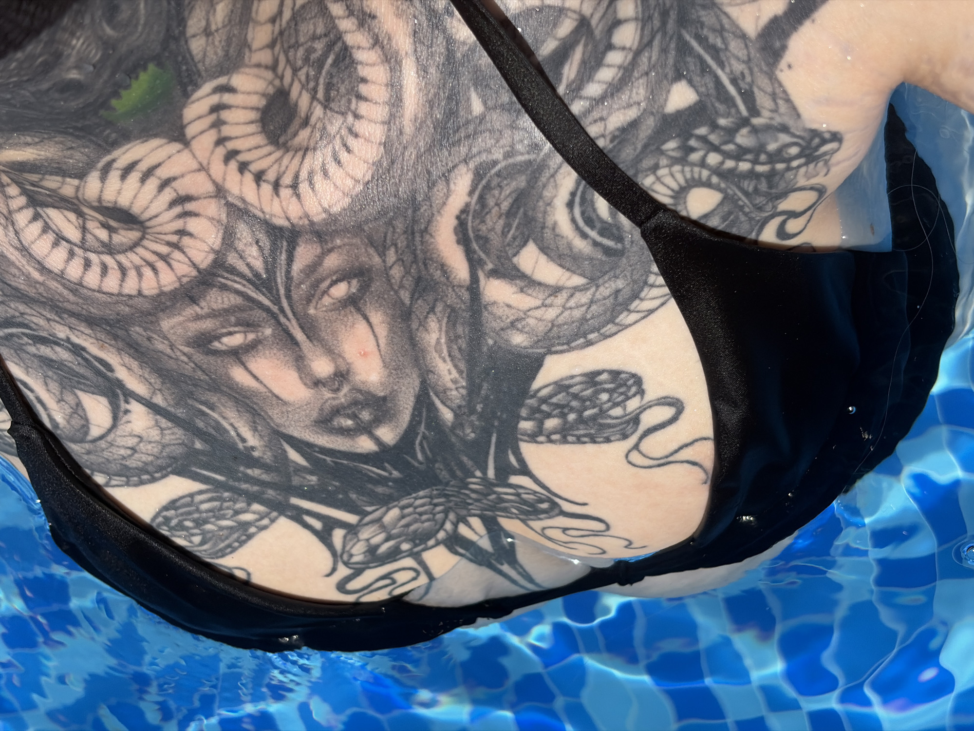 This media file captures a close-up view of a person's upper torso and chest, partially submerged in clear, vibrant blue water, likely in a swimming pool in Đà Nẵng, Vietnam. The individual is wearing a plain black bikini top.
The prominent subject is a large, detailed blackwork tattoo that covers much of the chest and extends towards the shoulders. The tattoo features a stylized, Medusa-like figure with a somewhat gaunt face, blank, eerie eyes, and numerous snakes coiling as her hair and around her. Several of the snakes are depicted with distinct scales and menacing heads. The tattoo appears wet, with small, shimmering glints of light reflecting off its surface and the surrounding skin, indicating water droplets.
The water itself is brightly lit and shows clear ripples and reflections of sunlight. The tiled pattern visible through the water's surface confirms it is a swimming pool. A small, green, leaf-like object is discernable floating near the top left of the tattooed figure's head. The bright illumination suggests it is daytime. The scene evokes a sense of relaxation or leisure by the water. There is no visible text in the image.