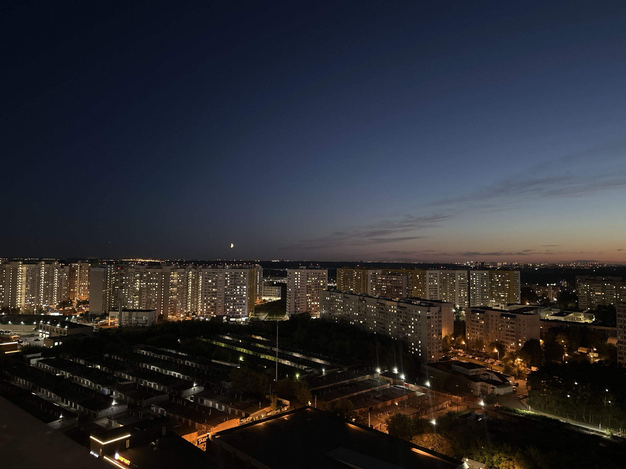 This is a nighttime aerial photograph of the city of Vidnoye, Russia. The image depicts a residential area with numerous apartment buildings illuminated by interior lights. In the foreground, rows of garages are visible, also lit by overhead lights. The sky is a deep blue, transitioning to lighter hues near the horizon, indicating dusk or twilight. A sliver of a crescent moon is visible in the upper left quadrant of the sky. The overall scene is one of a densely populated urban environment at night. There are no people or specific actions depicted in the image.