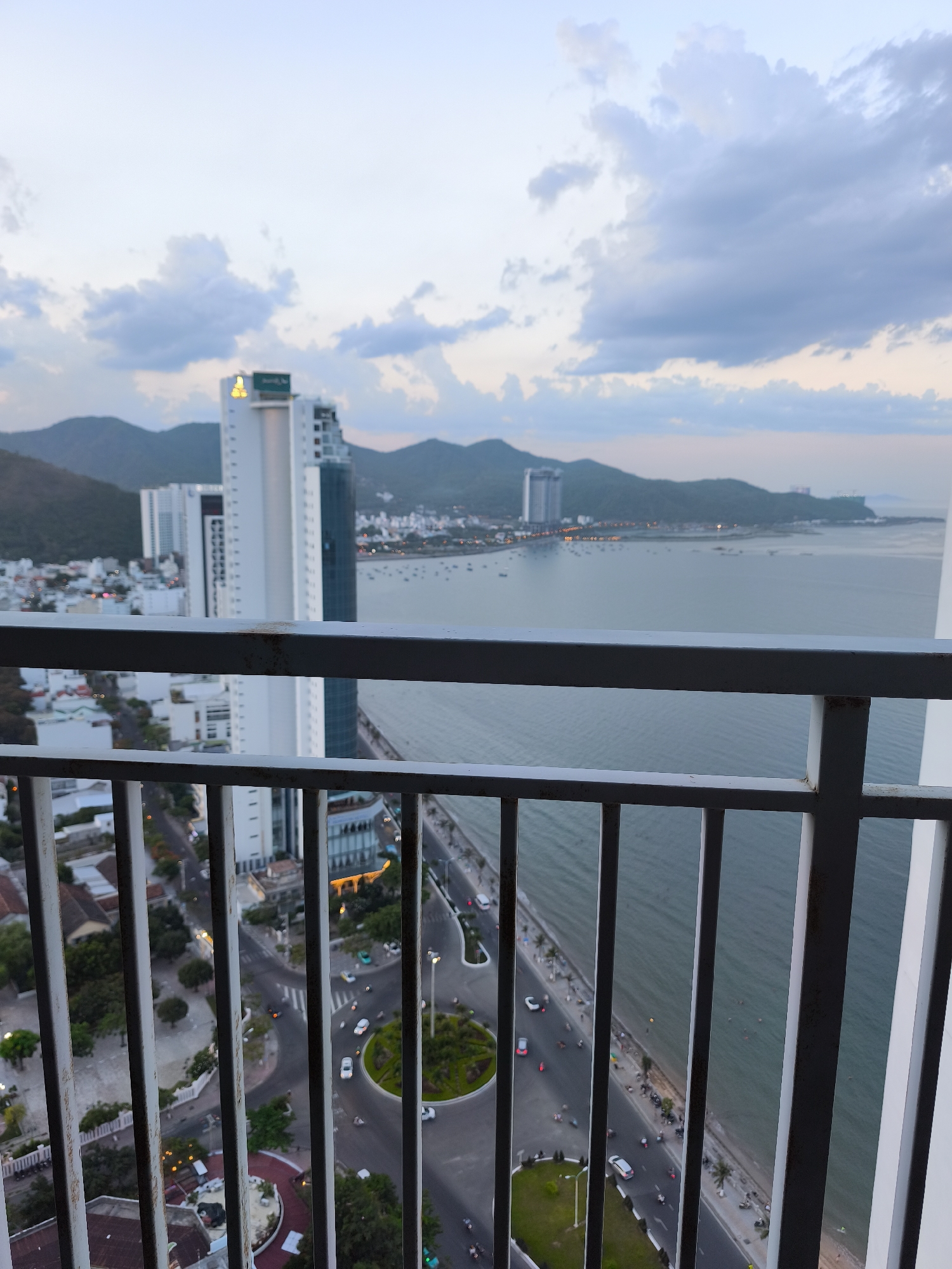 The image captures a panoramic view of a coastal city, likely Nha Trang, Vietnam, from a high vantage point. In the foreground, a white metal railing with vertical bars partially obstructs the view. Beyond the railing, a wide expanse of the ocean stretches to the horizon, where it meets a range of mountains under a cloudy sky.
The cityscape is characterized by numerous buildings, including several tall skyscrapers that dominate the skyline. One prominent skyscraper in the mid-ground is white with dark glass sections and is topped with a lighted sign. Along the coastline, a road runs parallel to the beach, with a steady flow of traffic consisting of cars and motorbikes. The beach itself appears to have scattered palm trees and a few individuals visible near the water's edge.
Further inland, smaller buildings and a network of streets can be observed, with roundabouts and streetlights indicating urban development. The overall scene suggests a late afternoon or early evening setting, with the sky exhibiting soft, diffused light. The perspective implies the photo was taken from a balcony or window of a tall building overlooking the city and the sea.